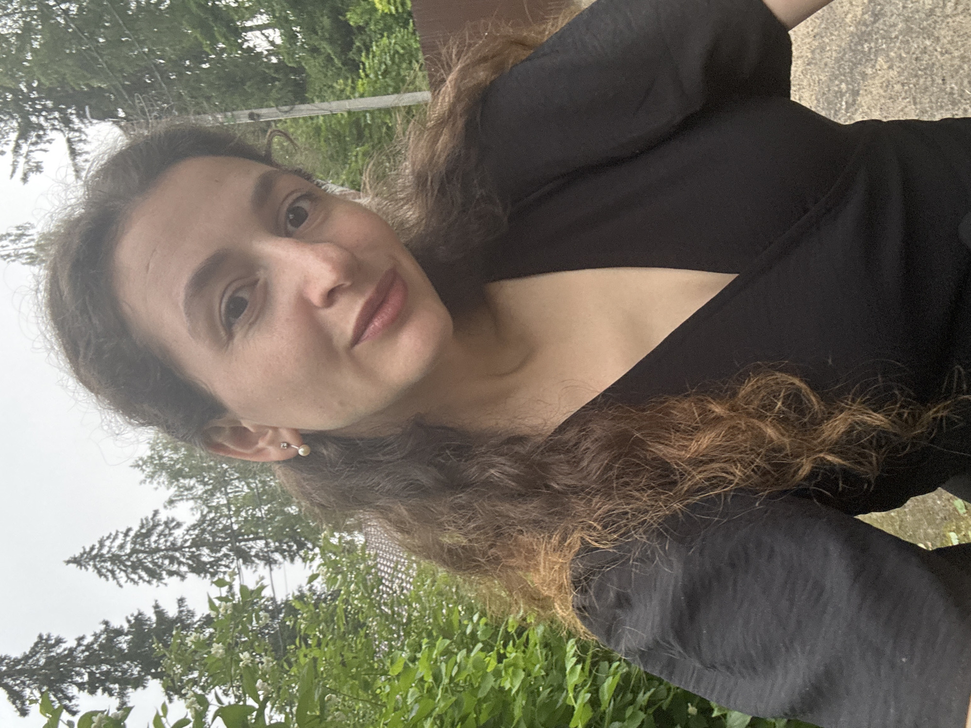 A medium shot shows a woman with long, curly brown hair wearing a black wrap top, standing outdoors. The woman is facing the camera, with her head slightly tilted. She has a neutral expression. The background consists of lush green foliage, tall trees, and a cloudy sky. A brown fence is visible on the right side of the image. The lighting suggests an overcast day.