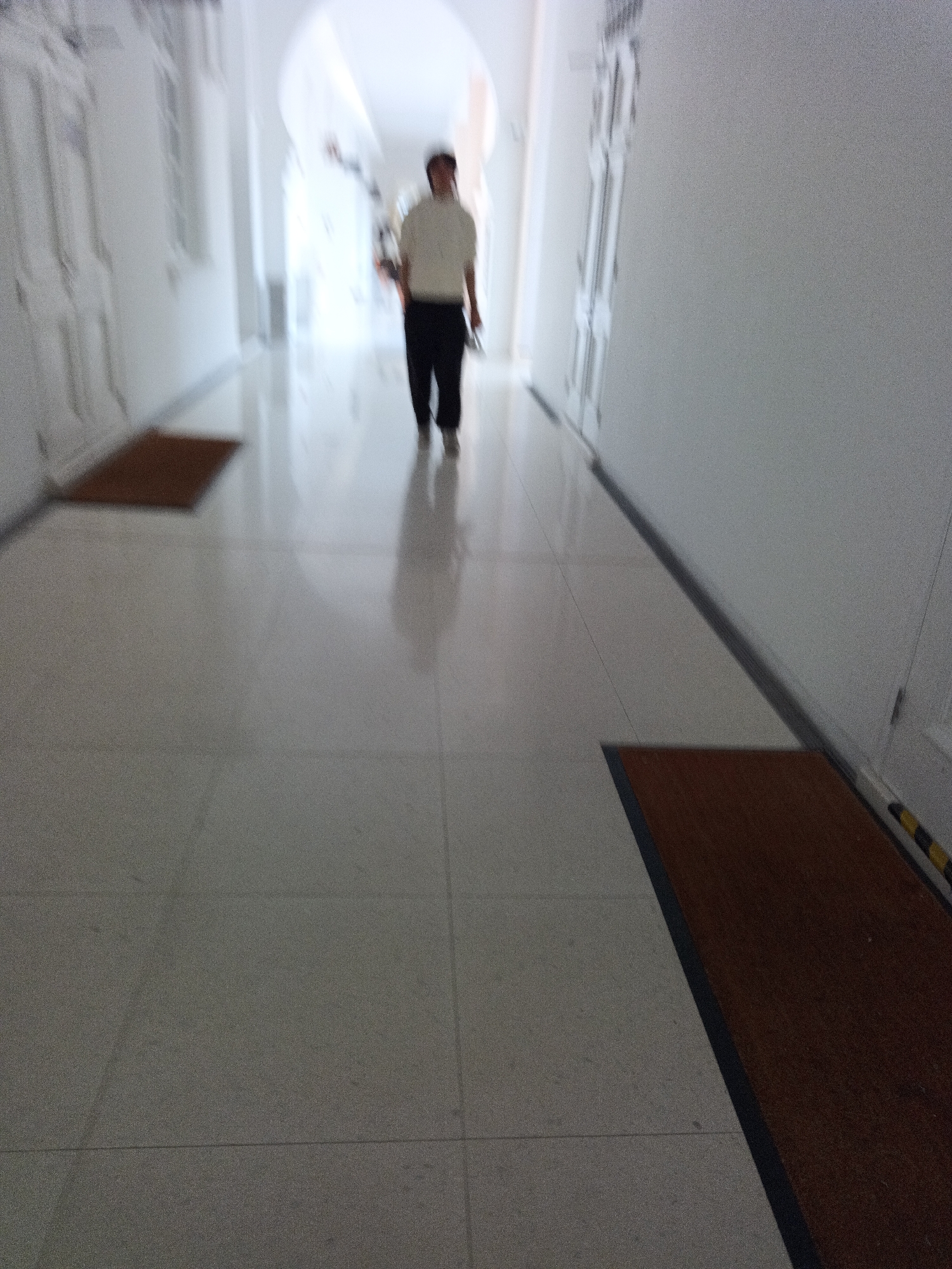 An outdoor, eye-level, full shot shows a person walking down a bright, tiled hallway towards the viewer. The person, who appears to be male, is wearing a light-colored collared shirt and dark pants. They are walking with their right arm slightly bent and holding a black object, possibly a phone, in their hand. The hallway is well-lit with natural light coming from an unseen source at the end of the hall. The floor is made of large, light-colored tiles that reflect the light, creating a glossy surface. Two brown mats are visible on the floor, one on the left side of the hallway closer to the foreground, and another on the right side further down the hall. The walls are white, and there are doors visible on the left side of the hallway. The image is slightly blurred, suggesting movement or a shallow depth of field. The scene is likely a public building or institution. It is difficult to determine the specific location within Kuala Lumpur, Malaysia, without further contextual information.