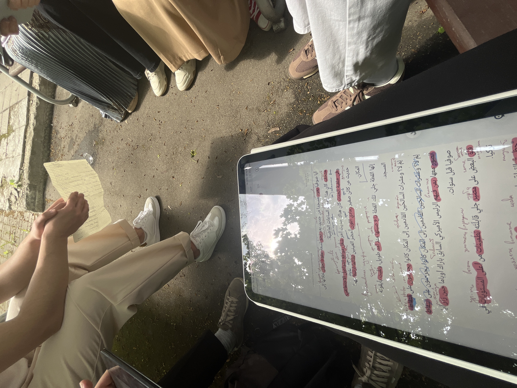 Here's a detailed description of the image:
**Scene Overview:** The image appears to be taken outdoors, possibly in a public space like a park or plaza, as suggested by the paving stones and trees visible in the reflection on the tablet screen. A group of people are gathered around a large tablet, which is displaying Arabic text with highlighted sections. The context suggests a possible discussion or study session related to the text.
**People:**
* Several individuals are present, but only their lower bodies and feet are visible. They wear a variety of clothing: pleated skirt, beige pants, and white sneakers.
* One person is holding a paper with writing on it.
**Objects:**
* **Tablet:** The central element is a large tablet displaying Arabic text with highlighting. Reflections on the screen obscure the text.
* **Paper:** Someone is holding a paper with what appears to be hand-written notes or text.
* **Clothing and Shoes:** These details indicate the individuals' personal style and attire.
**Actions and Interactions:**
* The people seem to be focused on the tablet, implying a shared activity like reading, studying, or discussing the information displayed on the screen.
**Environment:**
* The setting seems to be outdoors, with natural light and elements.
**Inference:**
Based on the text on the tablet, the highlighting, and the general appearance of the scene, it is likely that this is a study group or a discussion group. The location would be at an outdoor setting with pavement, trees, and natural light. The Arabic text relates to the Hagia Sophia museum in Turkey.
