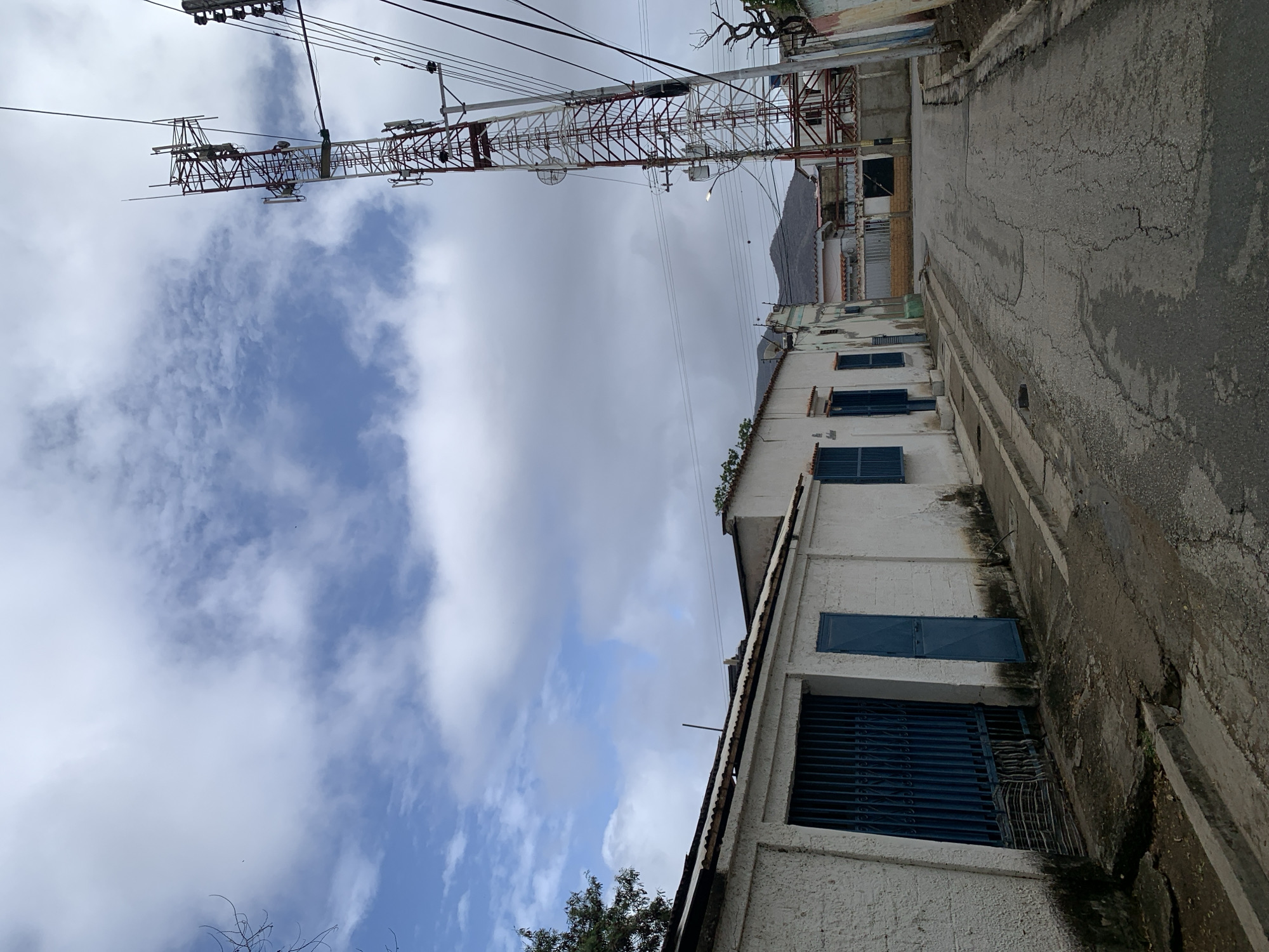An outdoor street scene in Santa Ana, Venezuela, is captured on a partly cloudy day. The perspective is angled slightly upwards, showcasing overhead elements.
On the left side of the frame, several white, stuccoed buildings line what appears to be a street or pathway. These buildings feature windows with distinct blue shutters and metal security grates. Above and behind these structures, a large, multi-tiered lattice-style communication or radio tower dominates the upper half of the image. The tower is constructed from red and white sections and is equipped with various antennas and dishes. Numerous power lines and utility cables are strung across the sky, connecting to poles and structures.
The sky is a mix of bright white and grey clouds, interspersed with patches of clear blue. In the distant background, faint outlines of mountains or hills can be seen. On the right, a cracked asphalt road runs alongside a concrete curb or drainage channel. Sparse tree branches and foliage are visible at the bottom and near some of the buildings.
The scene appears static, with no people, vehicles, or overt activity taking place. The overall impression is an ordinary view of an urban or semi-urban area, characterized by its utility infrastructure and local architecture. No visible text is present in the image.
