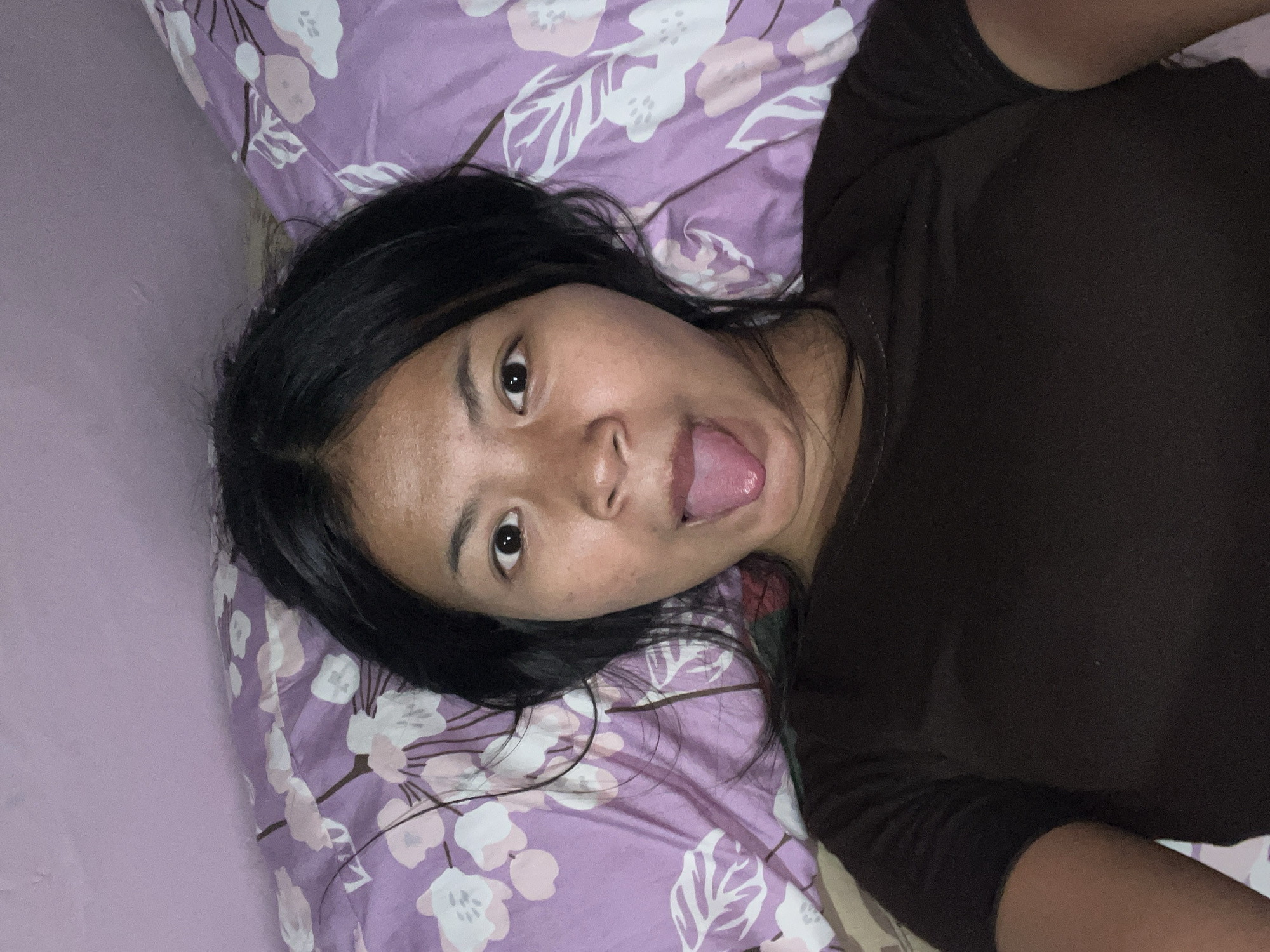 A young woman is lying in bed with her tongue sticking out, making a silly face. She is wearing a dark brown t-shirt. The bed is covered with a purple and white floral patterned sheet. The room appears to be dimly lit. The image is a selfie taken from a slightly elevated angle. It is not possible to determine the location in the city of Mendoyo, Indonesia from the image alone.
