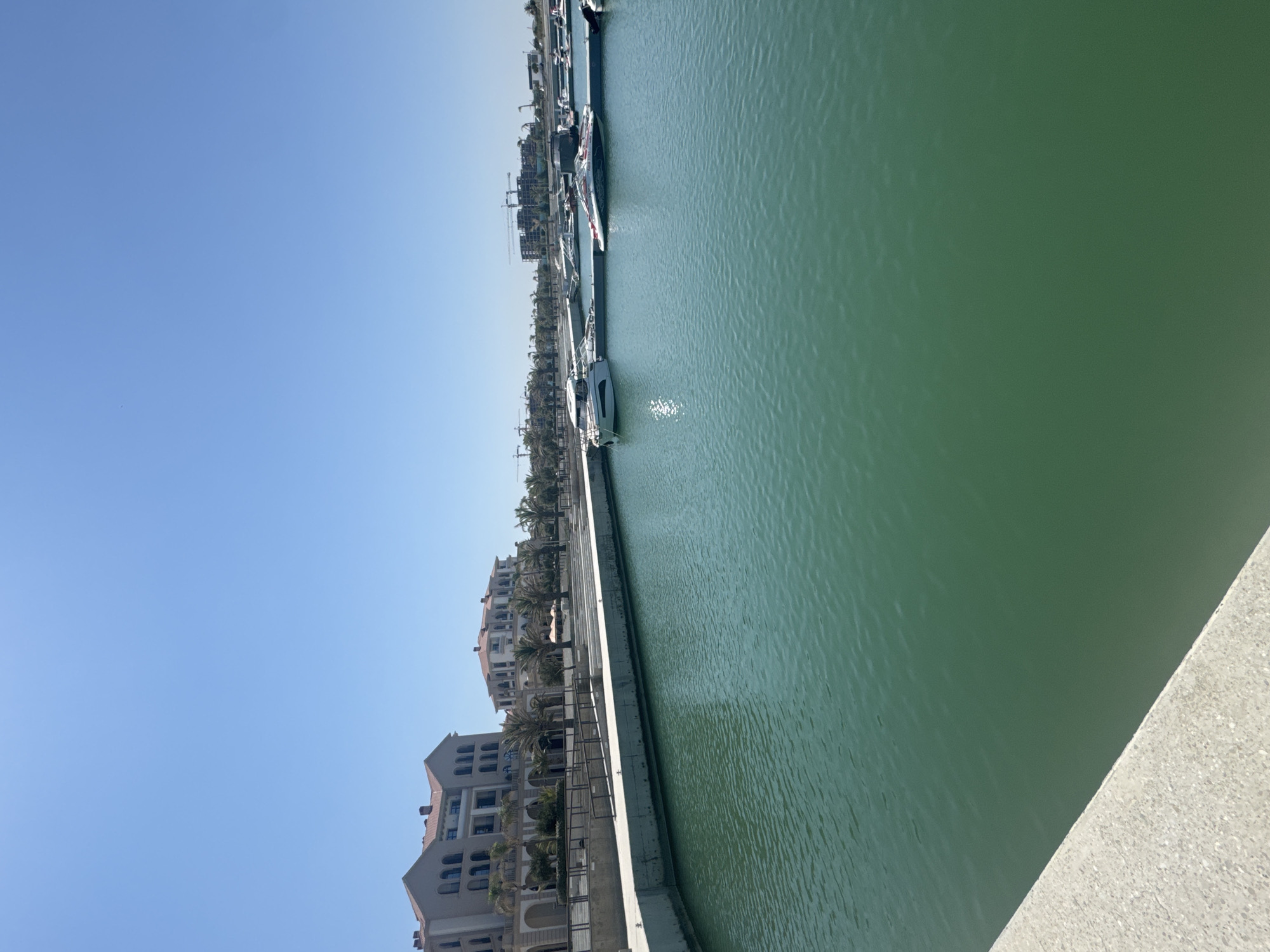 This image depicts a marina in Sabunchu Raion, Azerbaijan, on a clear, sunny day. The foreground is dominated by a calm, turquoise body of water, likely a section of the Caspian Sea. A concrete embankment with a light gray, textured surface lines the left side of the frame, leading the viewer's eye towards the scene. In the midground, several motorboats are docked along a pier. The boats vary in size and design, with some appearing sleek and modern.
Beyond the marina, a promenade runs along the waterfront, lined with palm trees. Behind the palm trees, a row of multi-story buildings with architectural elements reminiscent of Mediterranean design stands. These buildings feature light-colored facades, red tiled roofs, and numerous arched windows. Further in the background, construction cranes are visible, suggesting ongoing development in the area. The sky is a clear, bright blue with no clouds. The overall scene conveys a sense of leisure and development in a coastal urban setting.