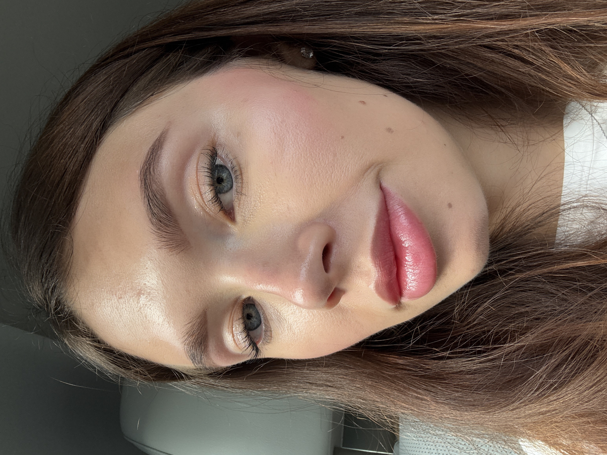 Close-up portrait of a young woman with light skin, brown hair, and blue eyes. She is wearing light makeup, including pink blush and lip gloss. Her expression is neutral, with a slight smile. The background is blurred and indistinct, but appears to be a neutral gray color. The location is not identifiable from the image.