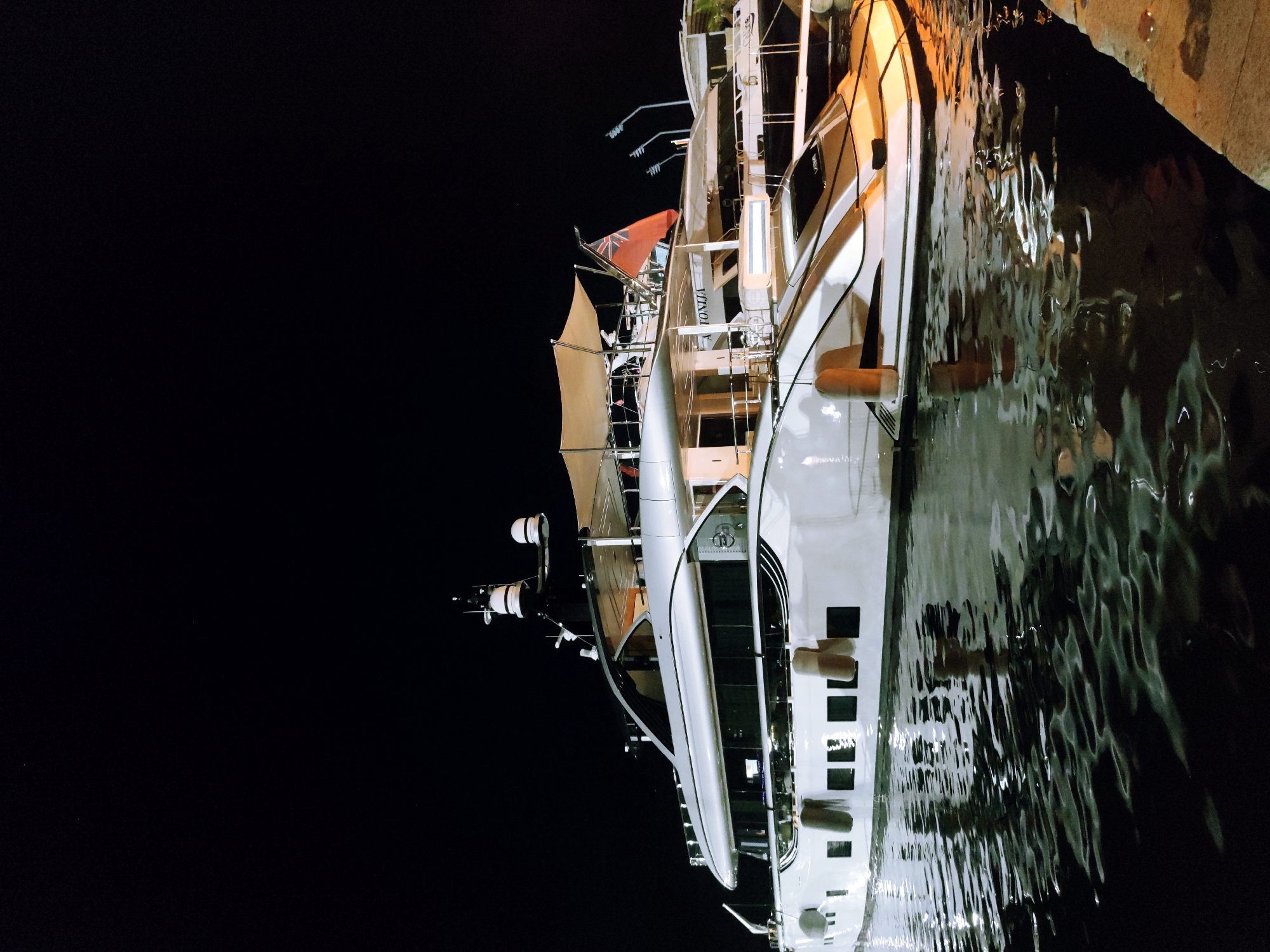 A large, white yacht is docked at night. The yacht has multiple decks and a red and white flag is visible on the top deck. The yacht is illuminated by lights on its exterior, which reflect on the water. The water is dark and choppy, with ripples and reflections. The background is black, indicating it is nighttime. The yacht appears to be in a marina or harbor, with a concrete dock visible on the right side of the image. The name "ANTONIA" is visible on the side of the yacht.