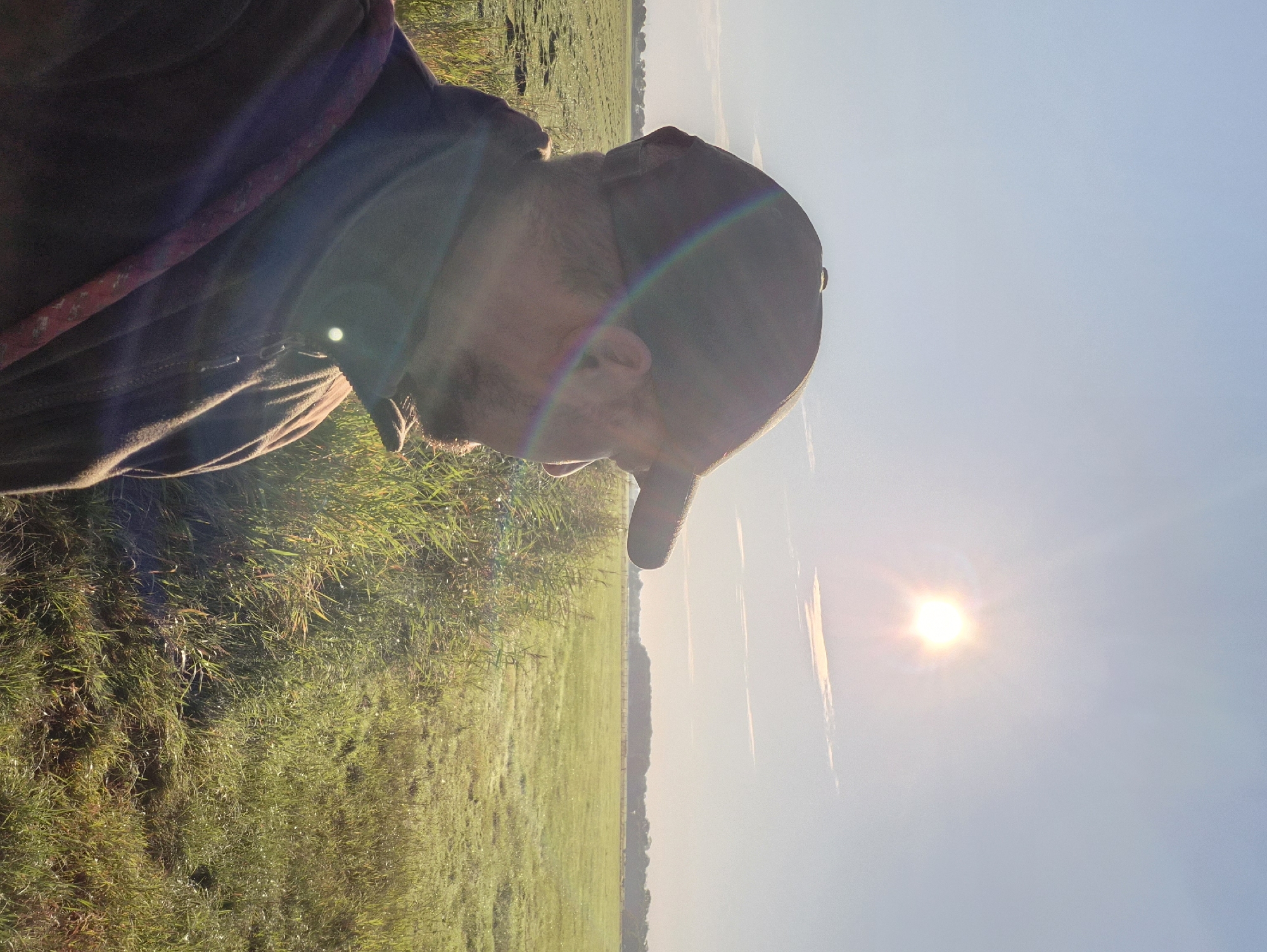 A man wearing a dark brown flat cap and a dark brown jacket stands in profile, facing right, in the foreground. He has a beard and is looking towards a sunlit, open field. The sun is high in the sky and creating a lens flare, with a rainbow-like effect visible on the man's cap. The field is green and appears to be covered in dew, suggesting it is early morning. Tall, green reeds are visible along the edge of the field in the foreground. In the distance, a tree line marks the horizon. The scene takes place in the city of Schiffdorf, Germany, specifically in a rural or agricultural area.