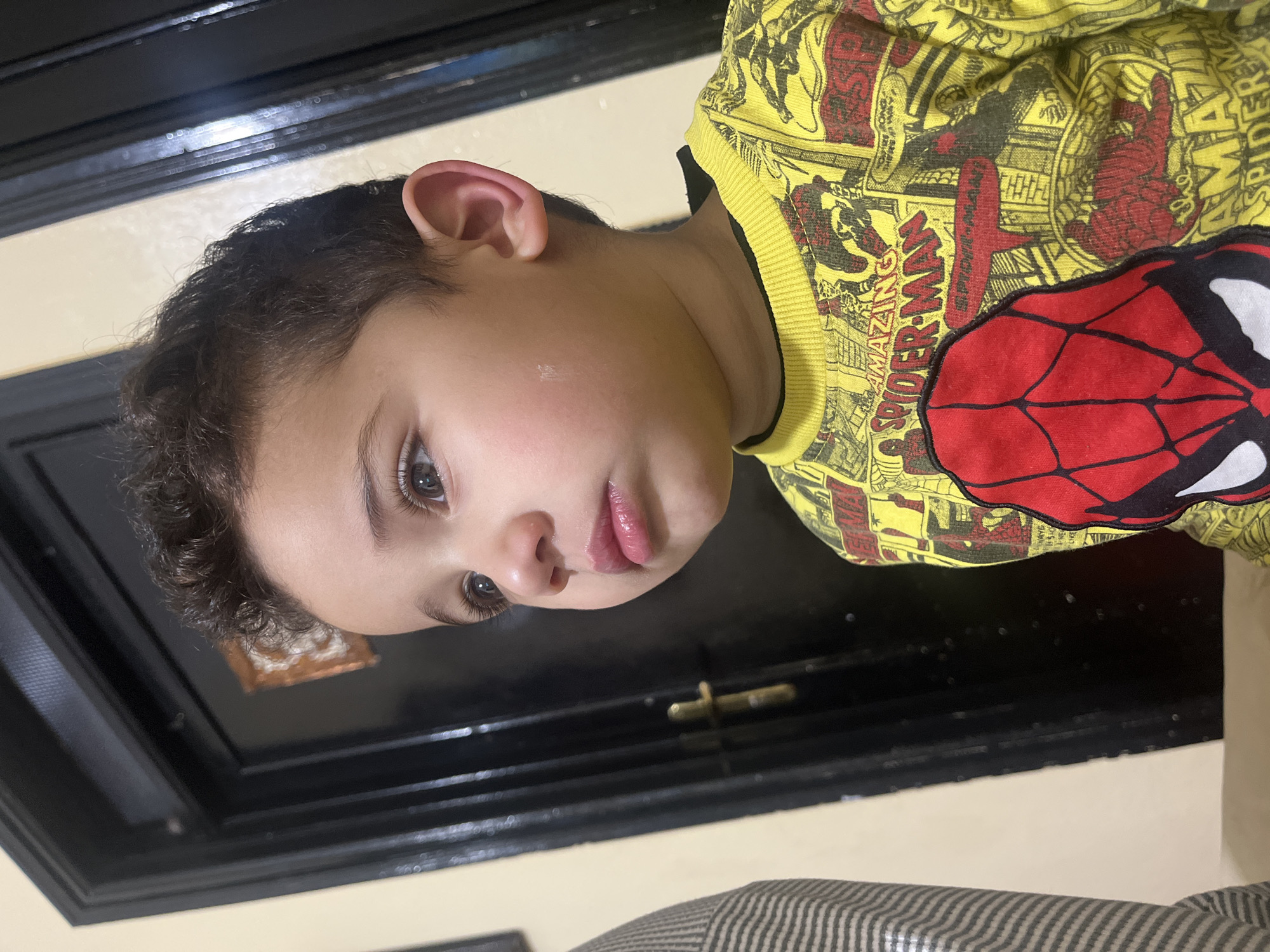 A young child wearing a yellow and red Spiderman sweatshirt is shown in a close-up, angled, indoor shot. The child has dark curly hair, fair skin, and light-colored eyes. A small, light-colored mark is visible on their cheek. Behind the child is a black object with a decorative metal handle, positioned against a beige-colored wall. A striped object is partially visible in the bottom right corner. The city of Ouaouizert, Morocco, is not discernible in the image.