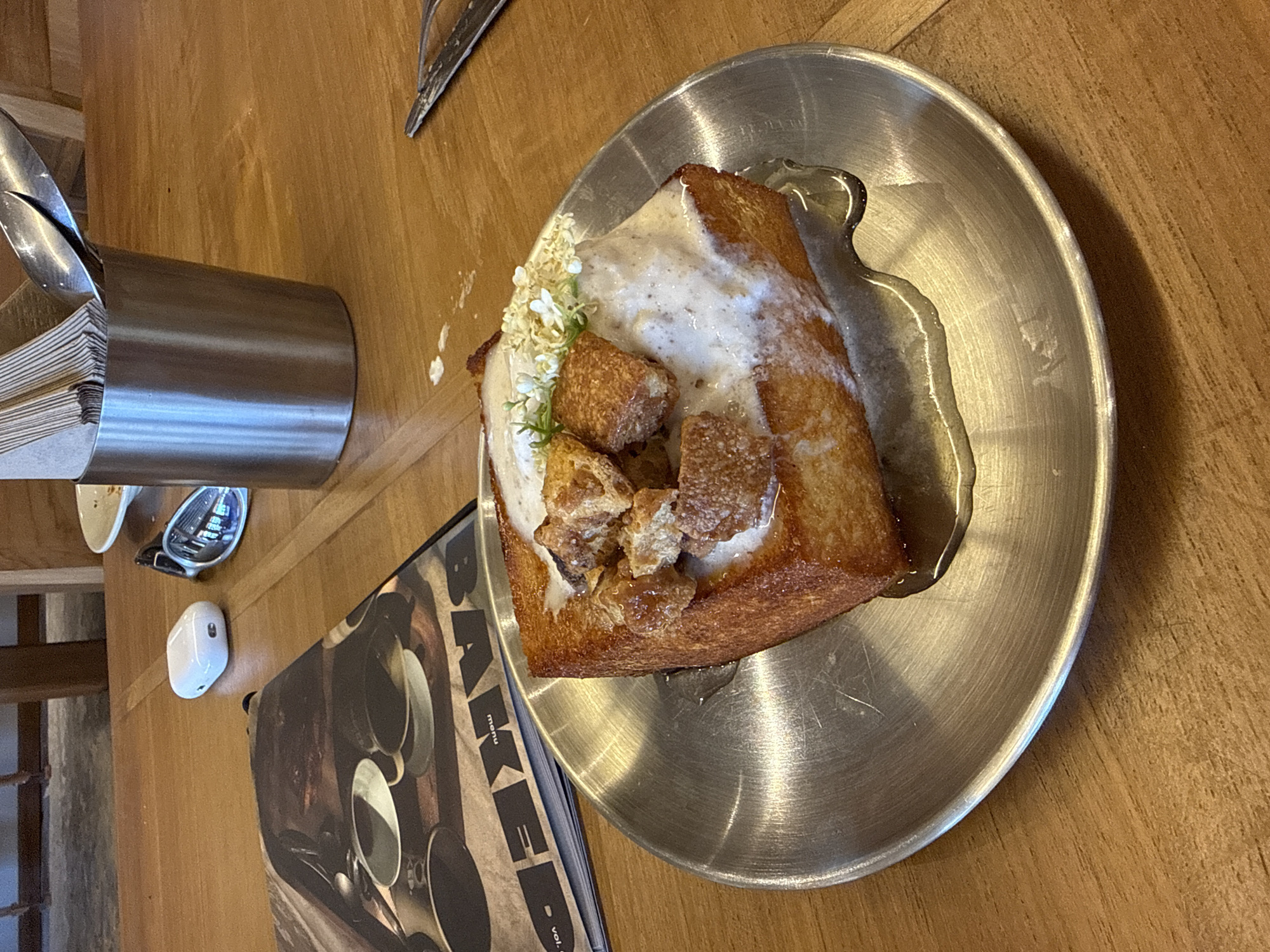 The image shows a wooden table with a silver metal plate holding a piece of thick-cut toast. The toast is topped with a white sauce, small fried croutons, and some small white flowers. A translucent syrup is visible around the toast on the plate. To the left of the plate, there is a silver metal container holding cutlery and paper napkins. A menu with the word "BAKED" printed on it is also visible. A pair of sunglasses and a white Apple AirPods charging case are also visible. The image was taken in North Kuta, Indonesia.