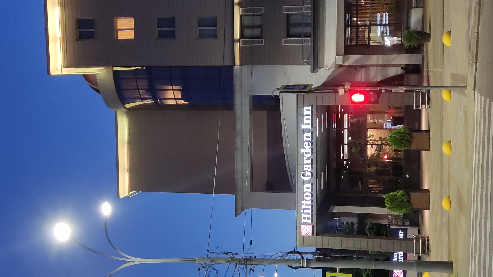 The image depicts the exterior of a Hilton Garden Inn hotel at dusk in Krasnodar, Russia. The building features a modern design with large windows and illuminated signage. The "Hilton Garden Inn" logo is prominently displayed in white lettering on a dark background, with the Hilton brand's distinctive pink flower emblem.
The scene is set on a street, indicated by a pedestrian crossing with white stripes and yellow spherical bollards. A traffic light displays a red signal. Streetlights with bright white bulbs are visible, suggesting it is either evening or early morning. The sky is a deep blue, characteristic of twilight.
No people are clearly visible in the frame, but the lit windows suggest occupancy within the hotel. The overall impression is one of a well-maintained and inviting hotel entrance. The presence of potted plants near the entrance adds a touch of greenery. The image is oriented vertically.