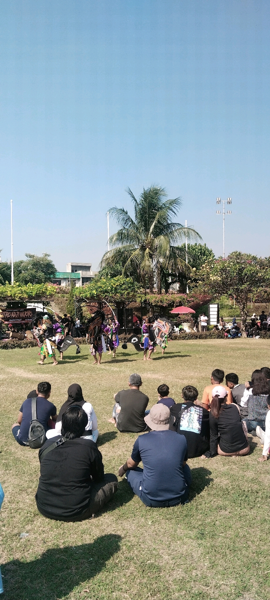 The image captures an outdoor cultural performance taking place on a grassy field under a clear blue sky. The performance features several individuals in elaborate costumes, some with headdresses and holding props, engaged in what appears to be a traditional dance or ritual. They are positioned on the grass in the mid-ground of the image.
In the foreground, a group of spectators is seated on the grass, facing the performance. The spectators are diverse in age and gender, dressed in casual attire. Some are looking directly at the performers, while others are conversing or looking elsewhere.
The background reveals more of the environment, including trees, some with lush foliage and flowers, and buildings that suggest an urban setting. Tall poles with lights are visible in the distance, indicating possibly a park or an event venue. A red umbrella is also visible among the trees. The overall scene suggests a sunny day and a community event. The presence of signage in the background, one of which reads "TRO ADJI WOYO," suggests the performance might be associated with a specific group or cultural event. This scene is consistent with the vibrant cultural activities often found in public spaces across Surabaya, Indonesia, where such outdoor performances are common.