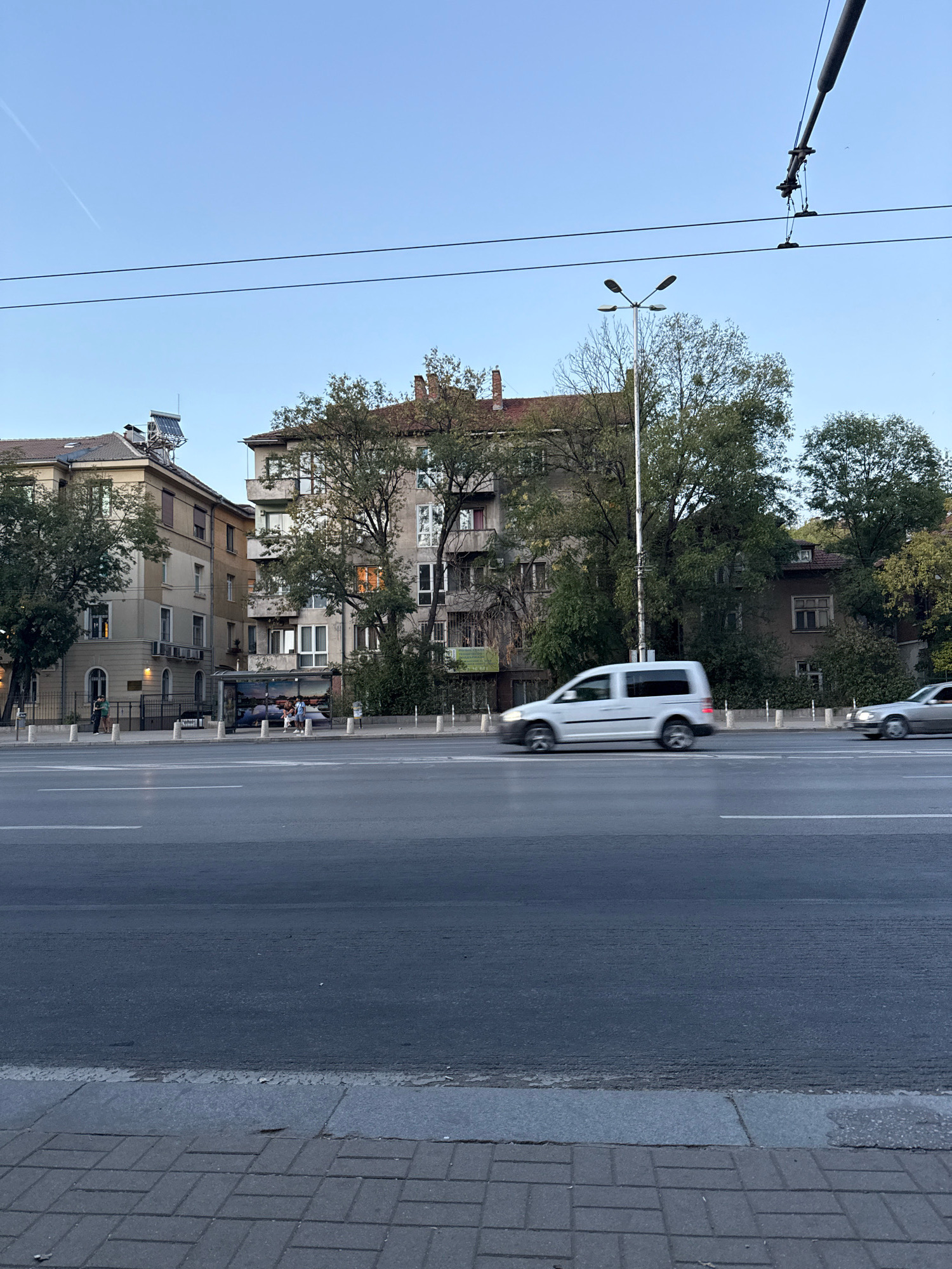 The image depicts a street scene in Sofia, Bulgaria, likely during the daytime or early evening, given the clear sky and the lighting. In the foreground, a paved sidewalk runs parallel to a wide asphalt road with visible lane markings. Several vehicles are in motion on the road; a white van is prominently in the center, captured with motion blur, and a silver sedan is visible further down the road, also blurred. Across the road, a row of multi-story buildings with architectural details suggestive of older European construction lines the street. Trees, some with full foliage and others with sparser branches, are interspersed between the buildings and along the sidewalk. A bus stop shelter is situated on the sidewalk, and a few individuals are present near it and near the entrance to one of the buildings. Overhead, electrical wires, possibly for public transportation, are strung across the sky, supported by poles. One pole has a street lamp attached. A solar water heater is visible on the roof of one of the buildings. The overall impression is a typical urban street in Sofia.