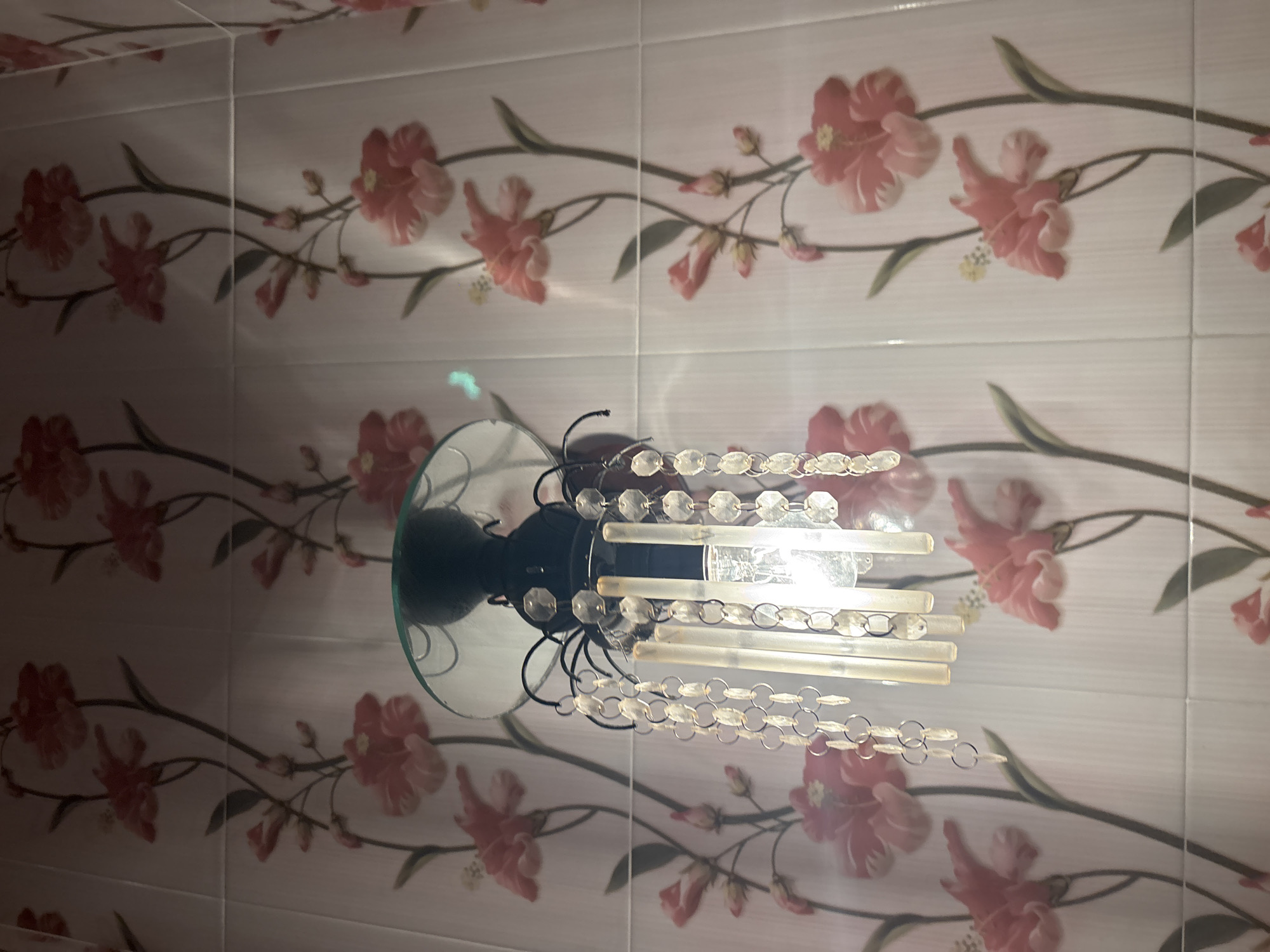 A close-up, eye-level shot shows a wall with a floral wallpaper pattern and a light fixture with hanging crystals. The wallpaper features a repeating design of pink flowers with dark green stems and leaves. The light fixture is black and has a glass shade. Dangling from the fixture are strings of clear, faceted crystals. The wall appears to be tiled. The image was taken in North Kuta, Indonesia.