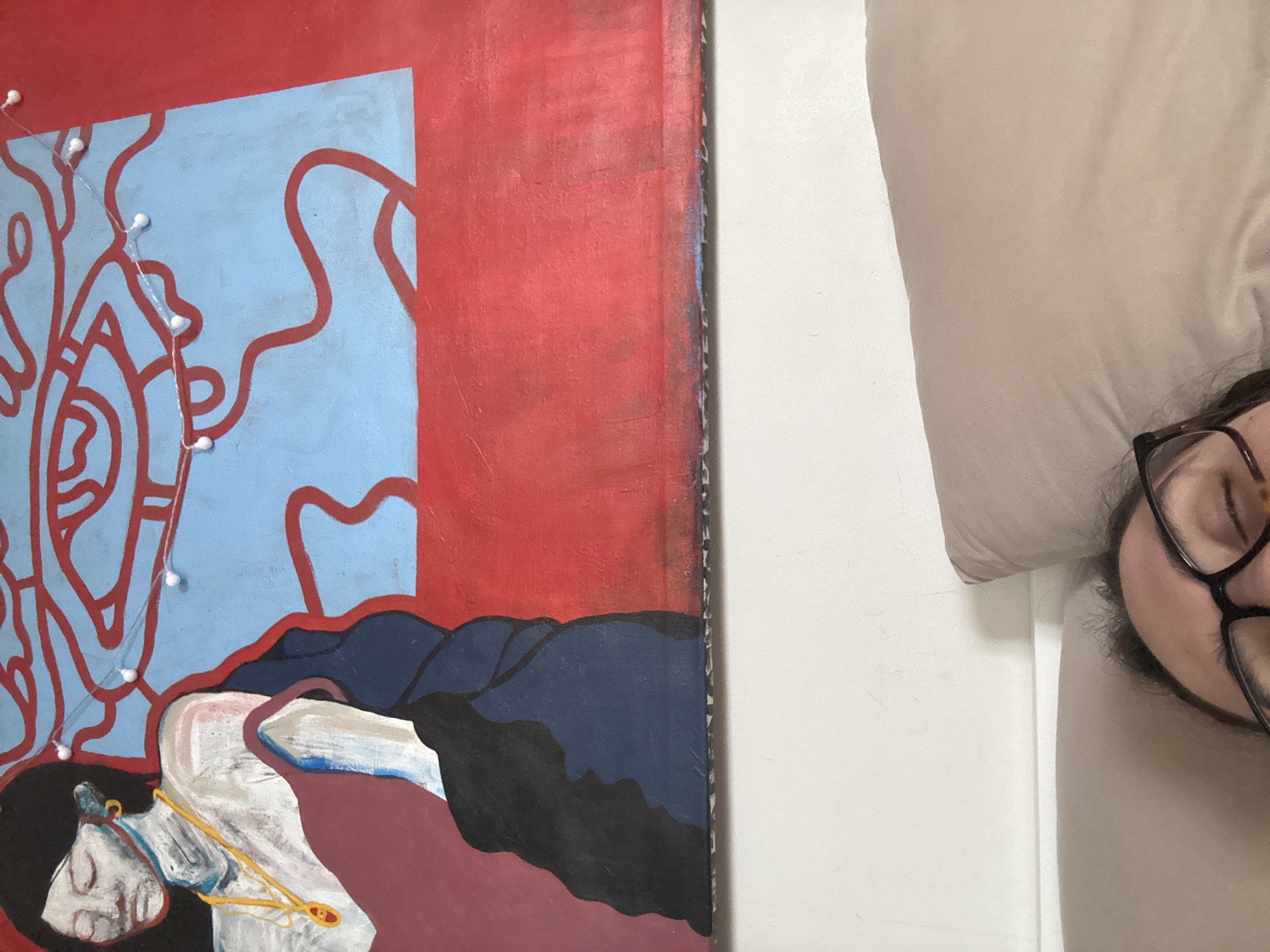 The image depicts a partially visible person lying on a bed with their head near a pillow. The person is wearing glasses. Next to the bed is a painting with a red background, a blue section with red line art, and a figure lying down. String lights are draped over the painting. The environment suggests an interior space. There is no contextual information to indicate that this scene is in Krasnoyarsk, Russia.