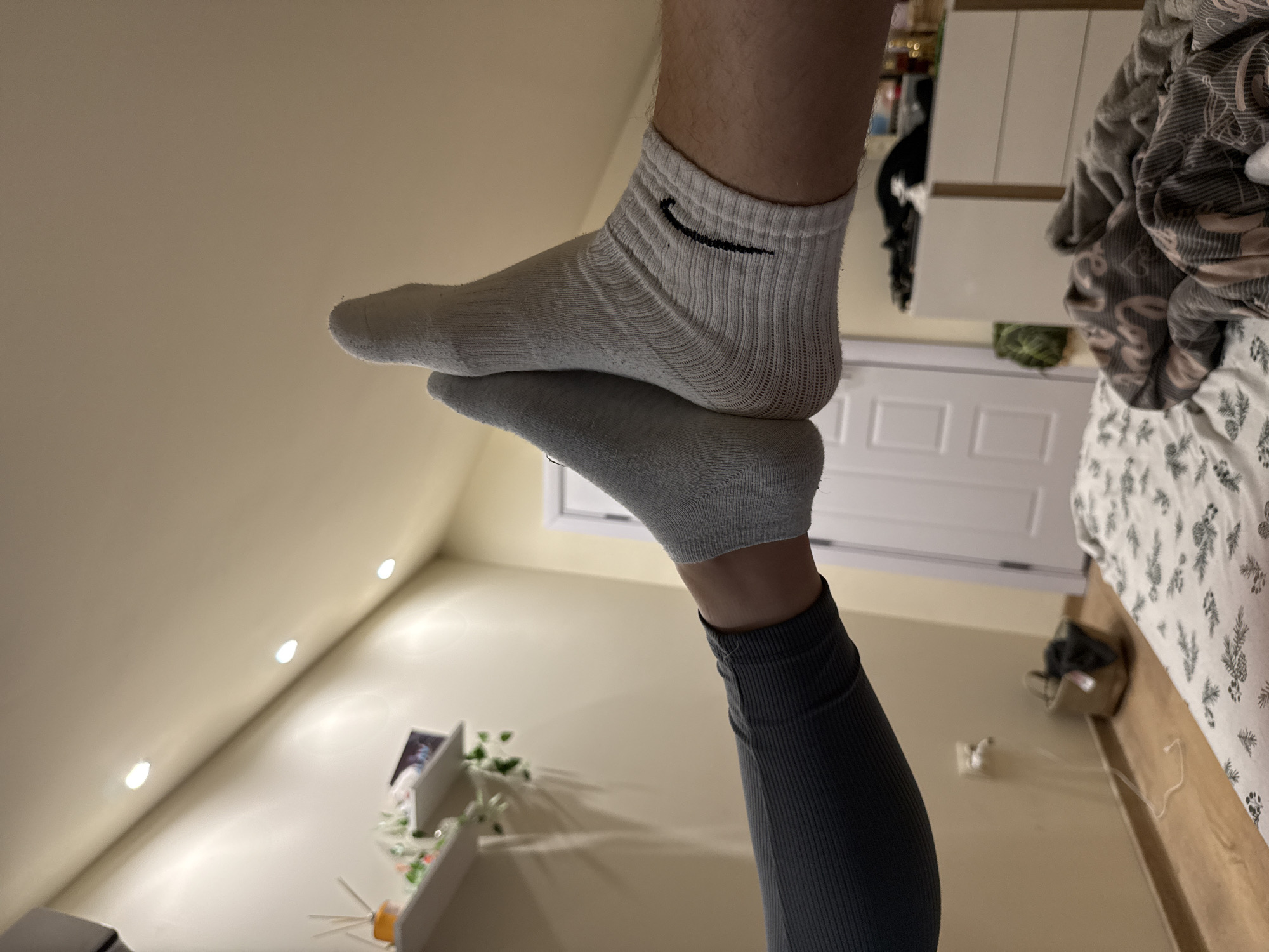 This image, taken in Dobrogled, Bulgaria, presents a low-angle, tilted view of two legs and feet in an indoor setting, likely a bedroom. The lower leg, wearing dark grey ribbed leggings and a grey ankle-length sock, extends upwards from the bottom of the frame. Resting horizontally on the toes of this foot is another bare leg, distinguished by its white ankle-length sock featuring a black Nike swoosh logo on the outer ankle.
The background reveals typical bedroom elements. To the right, a bed with a light wooden frame is partially visible, covered by a white duvet adorned with a pattern resembling green and grey pinecones or similar flora. Adjacent to the bed, a white chest of drawers or bedside table is present, along with a white electrical cable on the floor. Further back, a white paneled door is discernible. On the left side of the frame, several recessed spotlights are visible on the ceiling, casting a warm glow onto the light-colored walls. A wall shelf displaying a small green plant and other decorative items is also in view. The artificial lighting suggests an indoor setting, possibly during the evening. No explicit activity is depicted beyond the feet resting on each other in a static pose.