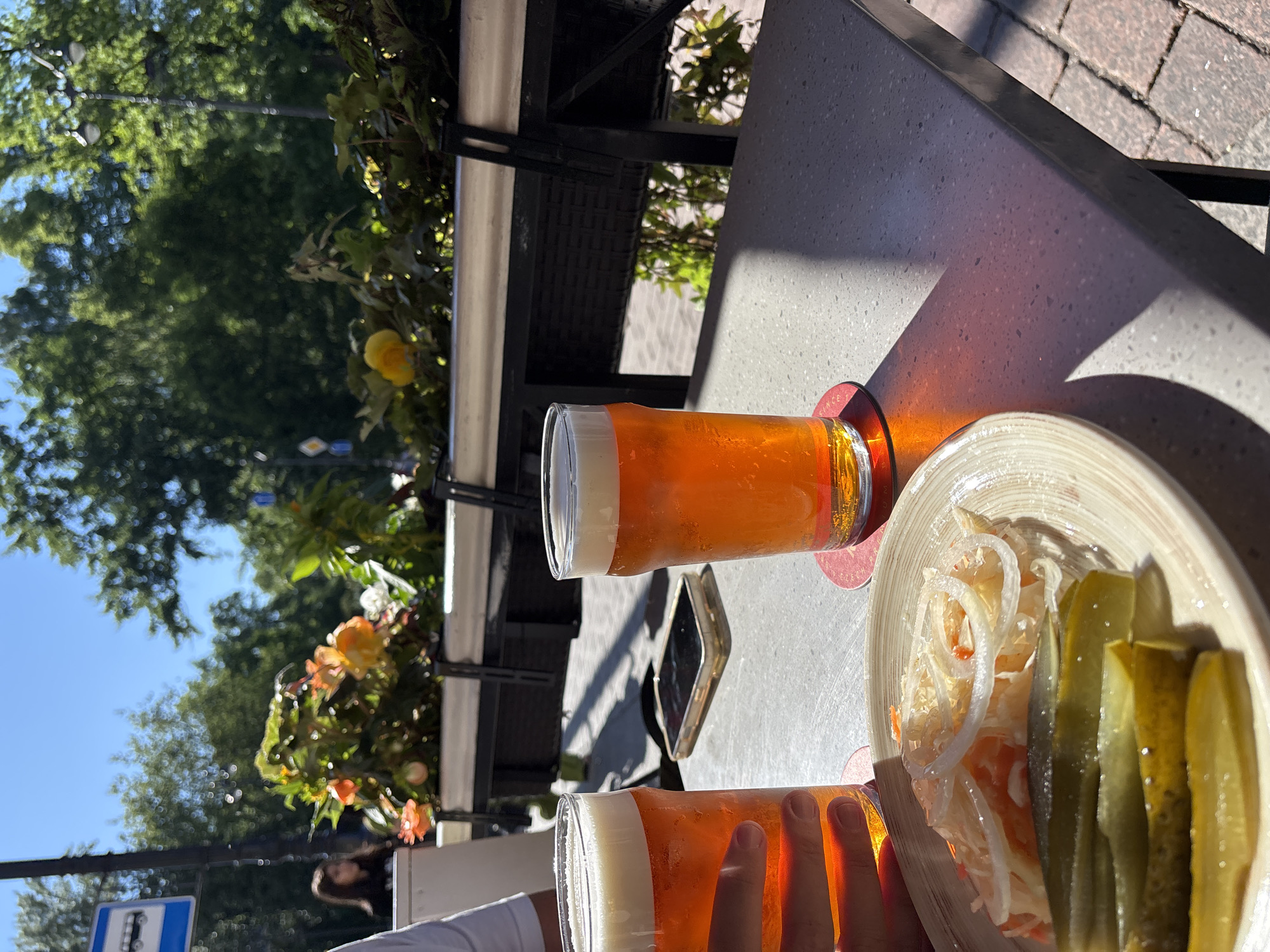 Two glasses of beer sit on a table outdoors. The beer is a bright orange color and topped with a thick white foam head. One glass is in the foreground, held by a person whose hand is visible. The other glass is slightly further back, to the left. A plate with a side dish of sauerkraut and pickles is to the right of the second glass. The table appears to be made of a light-colored, textured material, possibly granite or quartz. In the background, greenery from trees and bushes is visible, along with some orange and yellow flowers. The scene suggests a sunny day, with strong shadows cast on the table, indicating it is likely late morning or early afternoon. The presence of beer and a casual outdoor setting implies a leisurely activity, perhaps a meal or a break at a cafe. A sign with a symbol of a bus and a blue rectangle is visible on the left side of the image. The location context of Saint Petersburg, Russia, suggests this could be a common outdoor dining scene in the city during warmer months. No visible text besides the bus sign.