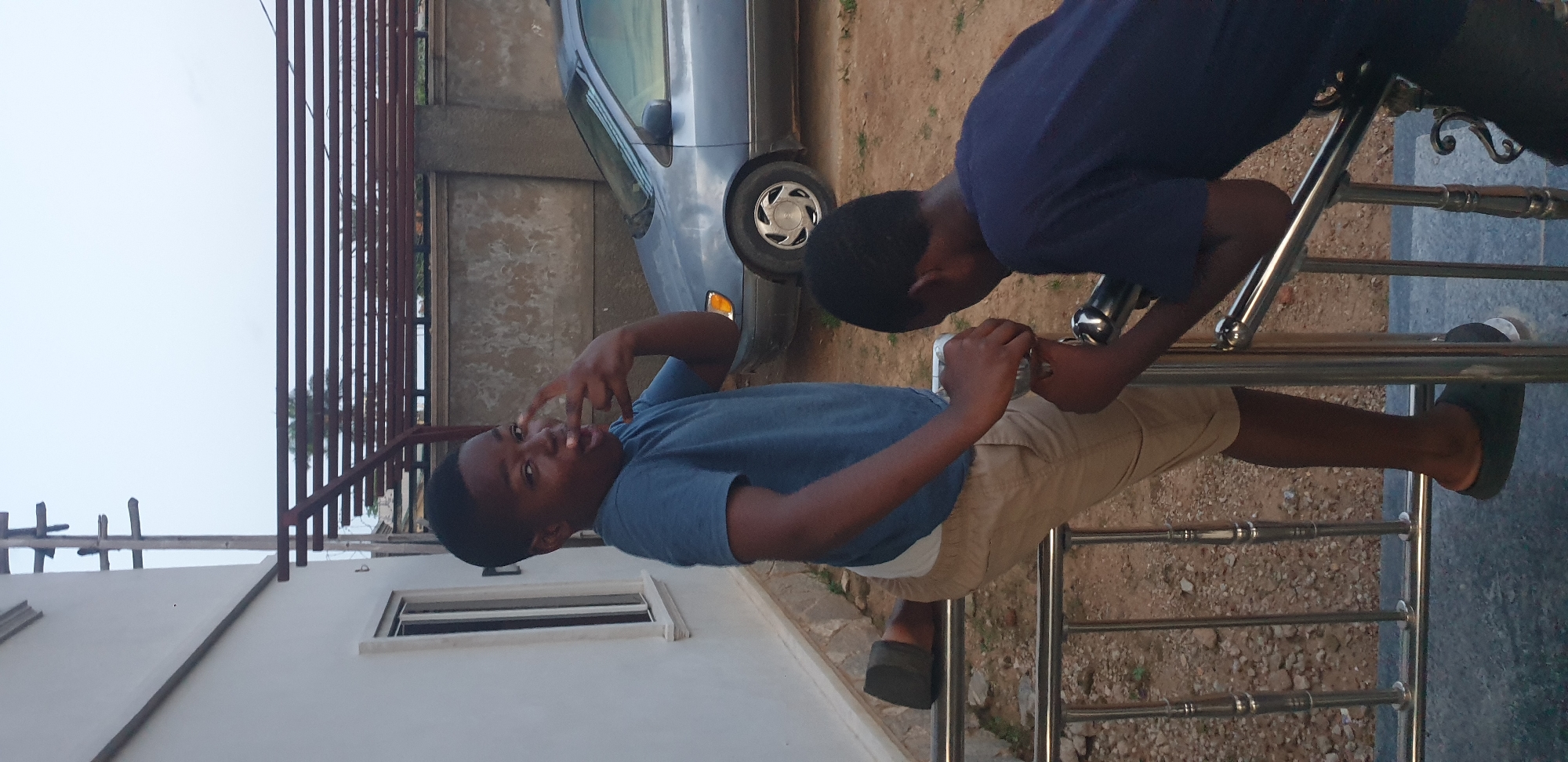 The image captures an outdoor scene in Narayi, Nigeria, featuring two young males near a residential structure and a parked vehicle. In the immediate foreground, a polished metal railing, likely part of a balcony or stairs, is prominent. Beneath the railing lies a dark grey, possibly tiled, surface, while beyond it, the ground consists of unpaved reddish-brown dirt and gravel.
On the left, a young male, likely a pre-teen, stands with his left foot resting on the railing and his right foot on the ground. He is dressed in a blue short-sleeved t-shirt, khaki shorts, and dark slip-on sandals. He faces the viewer, displaying an open-mouthed expression and forming a "peace sign" or "rock on" gesture with his left hand. His right hand is holding onto the railing.
To his right, partially obscured, a second, younger male in a dark blue short-sleeved t-shirt and dark trousers leans over the railing, looking downwards or away from the camera. Both of his hands are on the railing.
In the midground, a white building with a rectangular window is visible to the left. To the right, a light blue sedan car is parked on the dirt terrain, with its front passenger side and wheel visible. Behind the car, a textured concrete boundary wall extends horizontally. Above this wall, a dark metal structure, resembling a pergola or trellis with multiple horizontal bars, is present. Further in the background, a utility pole with cross arms stands against a pale, overcast sky. Some sparse, dark green foliage, possibly from a palm tree, is also discernible behind the metal structure.