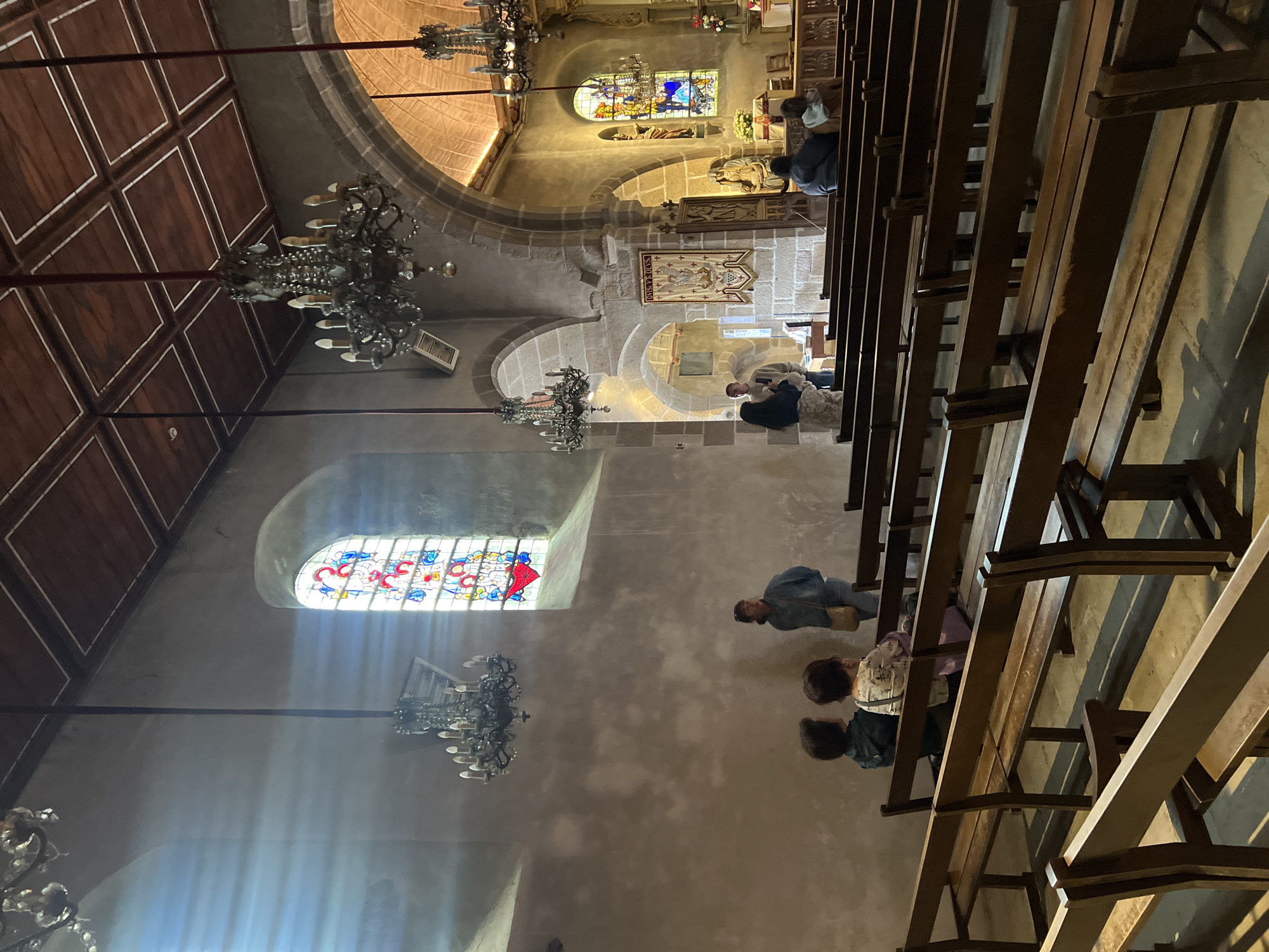 This image captures the interior of the Abbey Church of Mont Saint-Michel, located in the city of Le Mont-Saint-Michel, France. The perspective is from the nave, looking towards the chancel and altar area.
The scene is dominated by robust stone architecture, including weathered walls, large arched openings, and supporting pillars. A high ceiling features dark wooden coffered panels on the right side. Several ornate, multi-bulb chandeliers with metallic and glass elements hang from long dark rods at varying heights.
On the left wall, a tall, arched stained-glass window displays colorful religious figures, including a prominent red heart at its base. Sunlight streams through this window, creating distinct vertical light streaks on the adjacent plain stone wall and illuminated patterns on the floor.
In the foreground and midground, multiple rows of dark wooden pews with simple designs are arranged, facing the distant chancel. Several individuals are present in the space. On the left, two people are seated on a pew, with their backs to the viewer. Behind them, a woman stands, facing to the right, with a light-colored shoulder bag. On the right side of the nave and closer to the chancel, a small group of people is visible, some seated on pews, others standing.
The chancel area in the background-right features further stone arches, an altar setup, religious statues (including a light-colored statue resembling the Virgin Mary), and another stained-glass window. A rectangular banner, primarily red and white with religious figures and the Latin inscription "QVIS.VT.DEVS," hangs from a stone pillar on the right. A rectangular metal vent is visible high on a wall to the right. The floor appears to be stone or concrete, showing areas of light and shadow.