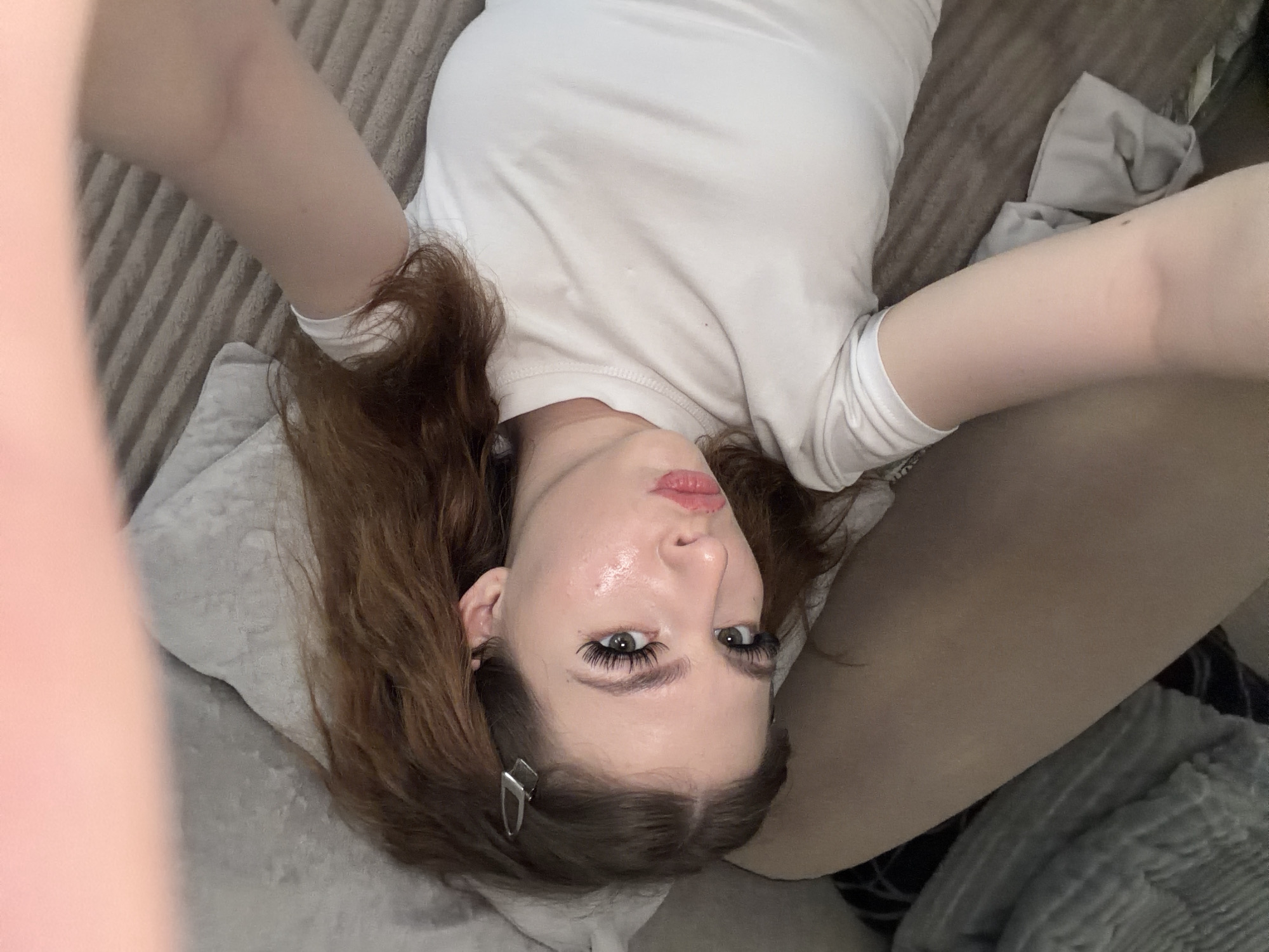 A young woman with auburn hair and fair skin is lying on her back on what appears to be a gray sofa or bed. She is wearing a white t-shirt. Her arms are raised above her head. A silver barrette is visible in her hair. The background includes textured upholstery and gray pillows or bedding. The scene is indoors. The location is not discernible from the image, and therefore cannot be identified as Noviy, Russia.