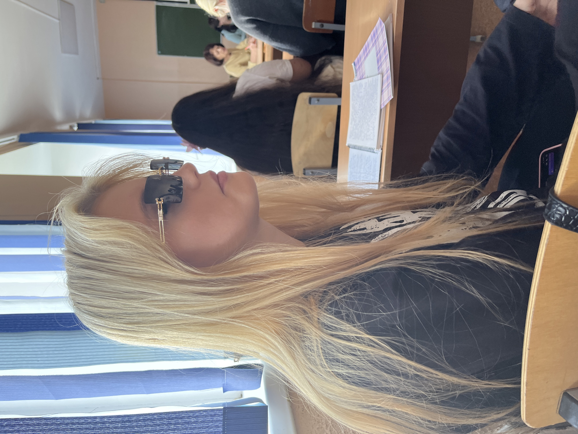 The image shows a young woman with long blonde hair wearing oversized black sunglasses and a black long-sleeved shirt. She is seated at a wooden desk in a classroom setting, with blue and white striped curtains visible behind her. Other students are seated at desks in the background. A notebook and papers are on the desk in front of her. The scene appears to be a classroom in Sterlitamak, Russia.