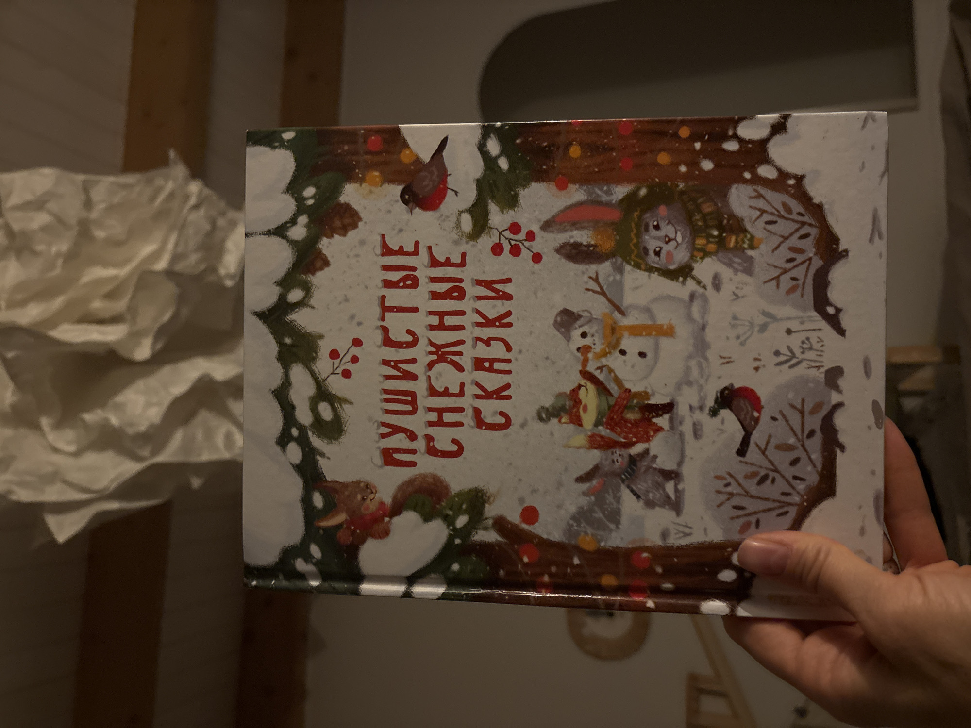 A person's hand holds a rectangular children's book, presenting its illustrated cover vertically. The cover art depicts a vibrant and whimsical winter scene featuring various animal characters in a snowy forest or park. A rabbit, wearing a green striped hat and scarf, stands near a cheerful snowman with a scarf and mittens. Another smaller snowman is visible nearby. To the left, a squirrel with a red scarf sits on a snow-laden branch, while a bullfinch with red markings perches above it. Another bullfinch and a badger-like animal are also part of the snowy landscape. The scene is framed by snow-covered trees and branches adorned with red berries and falling snowflakes, creating a festive, cozy atmosphere.
The book's title is prominently displayed in red Cyrillic script on the left side of the cover. It reads "ПУШИСТЫЕ СНЕЖНЫЕ СКАЗКИ," which translates to "Fluffy Snowy Tales" or "Furry Snowy Fairy Tales."
The image's background suggests an indoor setting with subdued lighting, indicating evening or nighttime. To the left, a crumpled, light-colored fabric object is partially visible, while wooden panels are seen further back, hinting at a home environment. The activity captured is someone holding the book, possibly for viewing or reading. The context provided indicates the location is Chaikovsky, Russia.