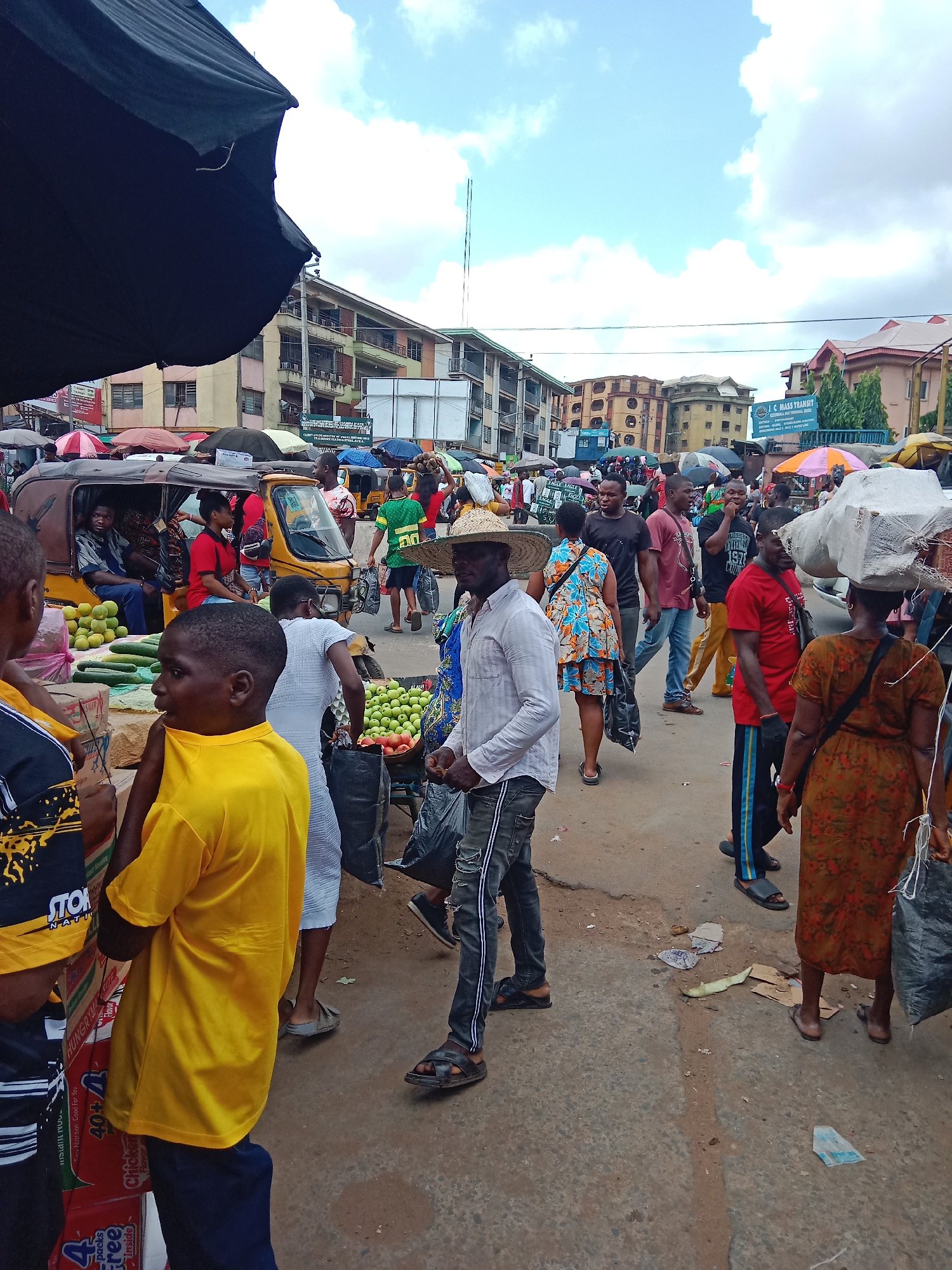 This image presents a bustling open-air market scene in Nkpor, Nigeria, under a partly cloudy daytime sky. The area is densely populated with numerous people—men, women, and children—engaged in various commercial activities.
In the foreground and midground, individuals walk through the crowded space. Many are dressed in casual attire; one person in a bright yellow t-shirt is prominent on the left. Market stalls display fresh produce, including green cucumbers, piles of limes or similar small green fruits, and red tomatoes. A yellow motorized tricycle, commonly known as a "keke napep," is visible with passengers, indicating local transport within the market. Large black umbrellas and colorful parasols provide shade over vendor areas. Several people are seen carrying goods, with one woman balancing a large, wrapped bundle on her head.
The background features multi-story buildings with balconies, characteristic of urban Nigerian architecture. A large signboard reads "C MASS TRANSIT," likely indicating a transport service. The ground is paved but appears dirty with scattered debris, reflecting the constant activity.
Visible text from packaging includes phrases such as "4 packs Free," "Nutrition, Good For You," "40+ Inside Chicken Flavor," and the word "HUNGRYUE." The year "1977" is also partially visible on an object. The overall atmosphere conveys a lively and active commercial hub, typical of a busy African market.