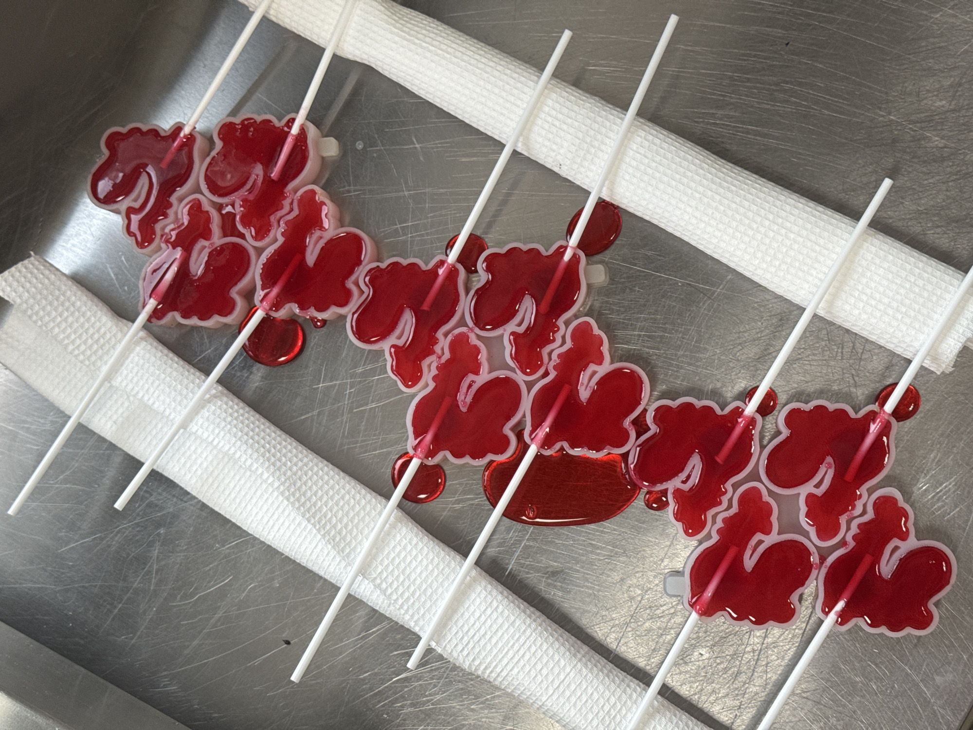The image shows a metal tray with a row of red, candy lollipops shaped like Cyrillic letters with white plastic sticks. The lollipops are resting on two rolled white towels. The setting appears to be a commercial kitchen or food preparation area due to the stainless steel tray and professional, sanitized appearance. There is no evidence indicating the specific location of Perm, Russia.