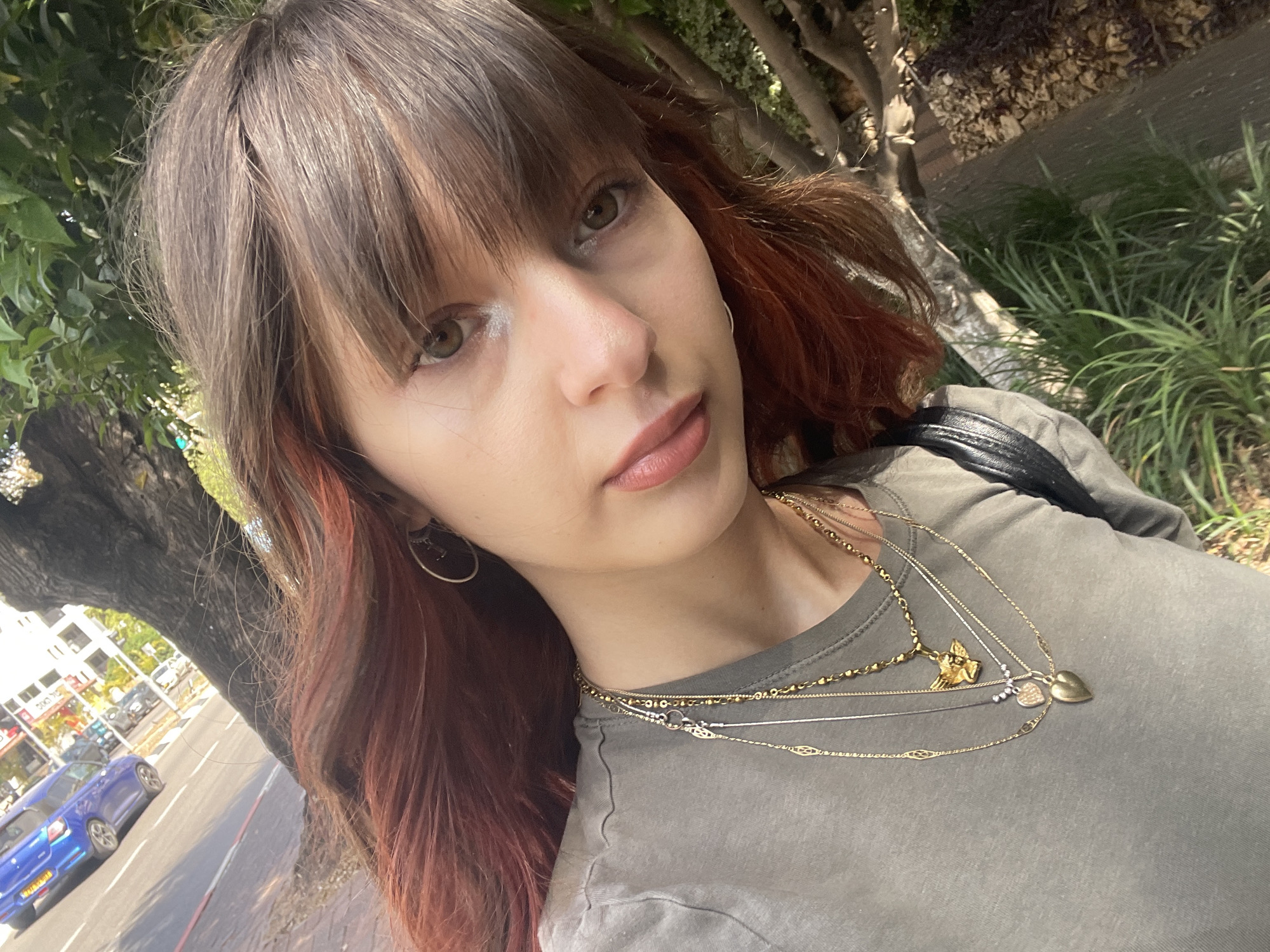 A medium-shot selfie photograph shows a young woman with shoulder-length brown hair and reddish-brown highlights. She has fair skin, light eyes, and is wearing makeup. She is wearing a grey-green t-shirt and several layered gold necklaces with different charms. She has a silver hoop earring in her left ear and a black shoulder strap over her right shoulder.
The background shows an urban street scene, with trees and greenery to the left and right. A street with cars is visible in the background, and buildings can be seen across the street. Based on visual clues and the prompt, the location is Raanana, Israel.