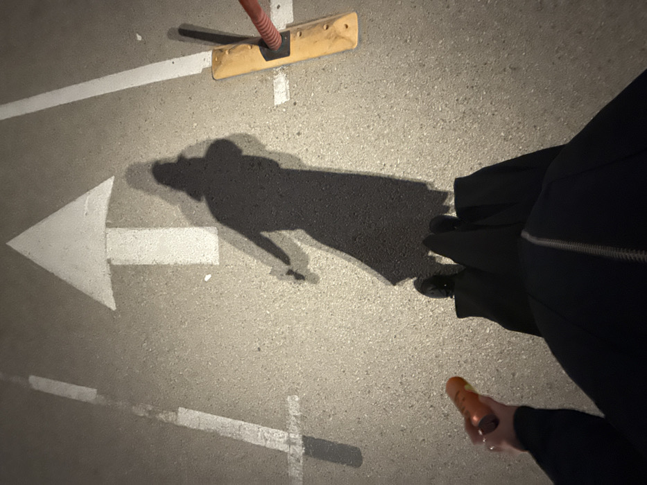 An overhead view shows an expanse of gray asphalt marked with white directional arrows and lane lines. A shadow of a person wearing a long dark coat falls across the center, obscuring part of a left-pointing arrow. The person holds a small brown object. A wooden broom with an orange handle sits on the upper part of the frame, casting its own shadow. The overall lighting suggests an evening or nighttime setting.