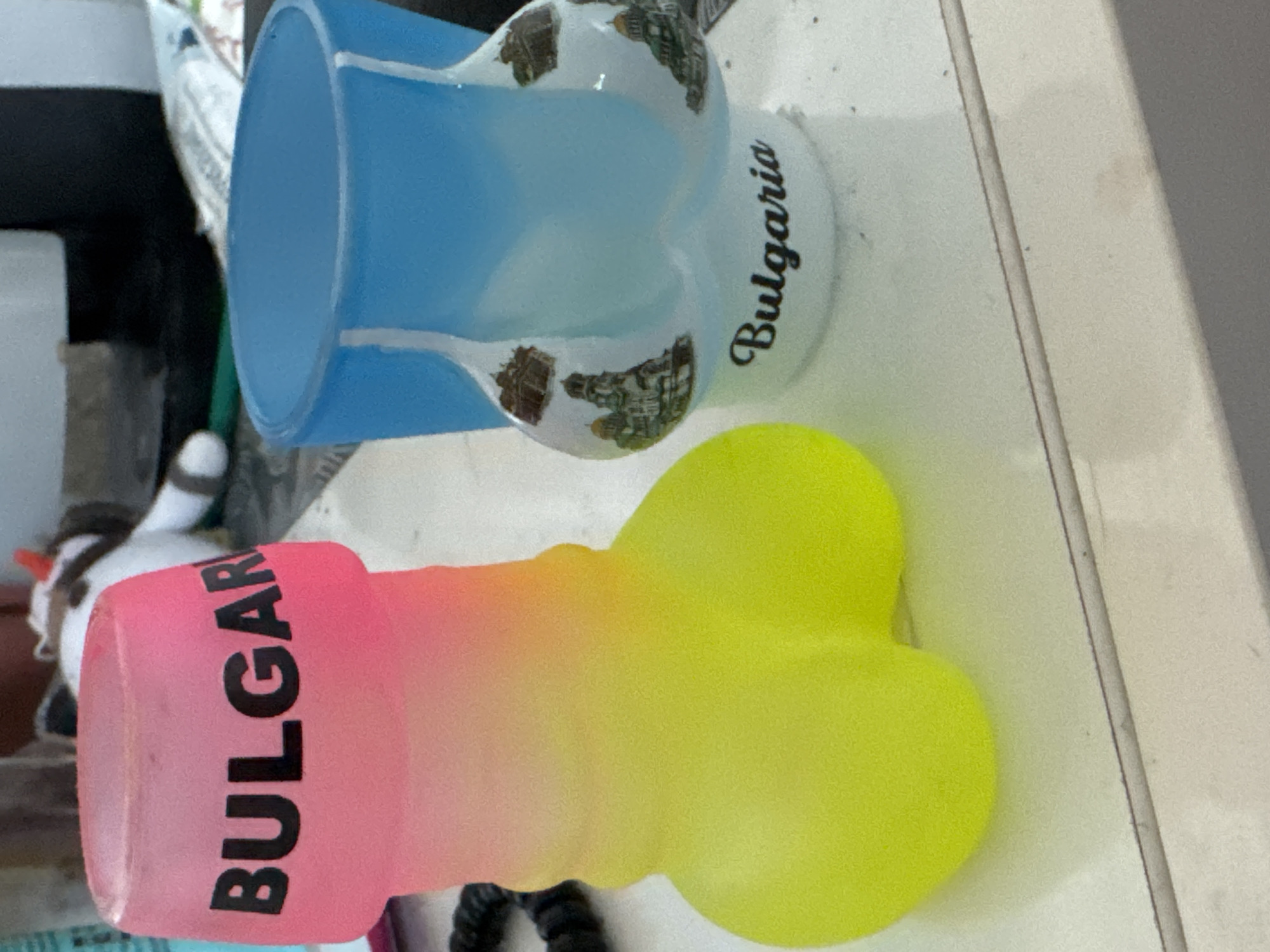 Two phallic-shaped shot glasses are displayed on a white surface, likely a table or shelf. The shot glass on the left is pink and yellow, with the word "BULGARIA" printed in black letters. The shot glass on the right is blue and white, featuring images of architectural landmarks and the word "Bulgaria" written in script. The background is blurred but suggests an indoor setting with other objects, including a stuffed animal. The image provides no information about the city of Sofia or any specific location.