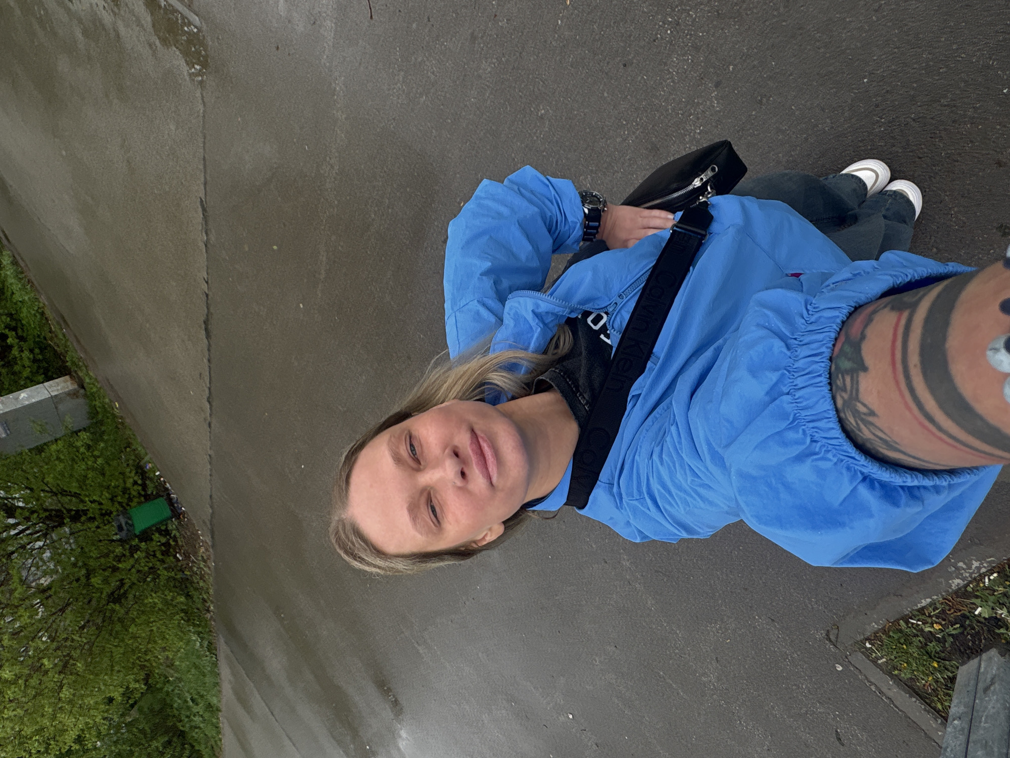 A person with fair hair stands on a wet, gray concrete surface, possibly a sidewalk or path. The individual is wearing a light blue jacket over a black shirt with the word "Calvin Klein" visible on the strap of a black shoulder bag. They have jeans and white sneakers on. A tattoo is visible on the person's right arm. In the background, there is some greenery, potentially trees or bushes. Based on the image, the specific location in Perm, Russia cannot be determined.