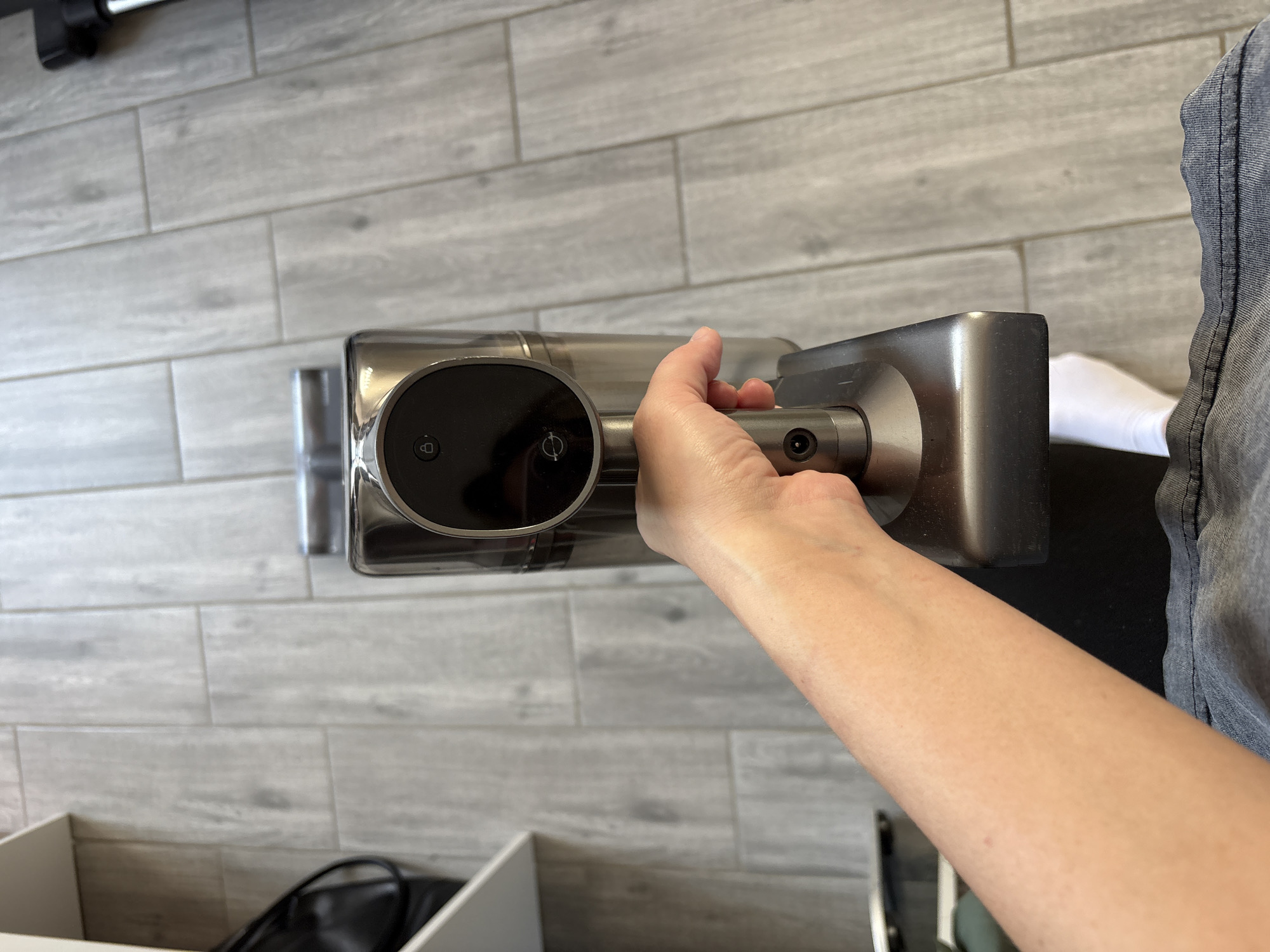 The scene, located in Votkinsk, Russia, is depicted from an elevated perspective looking downward onto an indoor floor. A fair-skinned human right hand, with visible knuckles, grasps the metallic-grey handle of a cordless stick vacuum cleaner. The vacuum's main body, also metallic-grey, features a translucent dust collection bin and a dark control panel exhibiting two illuminated white icons: an open padlock symbol positioned above a swirling, propeller-like symbol. A circular port is present on the vacuum's body below the control panel. The lower portion of the image reveals the person's forearm and lap, clad in grey denim fabric with prominent stitching, and a segment of a white sock on the right foot. The floor is composed of light-grey, rectangular planks or tiles arranged in a staggered pattern, mimicking a wood grain texture. In the upper-left background, a segment of white shelving is visible next to a partially obscured black bag. To the upper-right, a black and silver component, consistent with a vacuum attachment, is partially discernible.