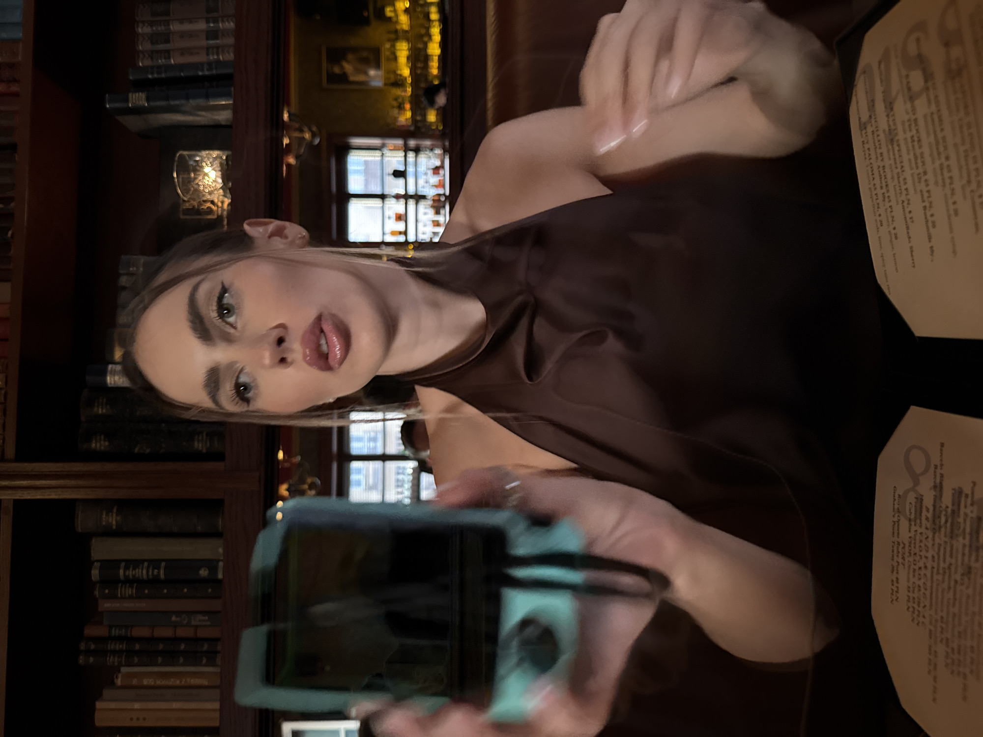 A young woman is holding a phone in front of her face, presumably taking a selfie or video. She is sitting at a table in a dimly lit establishment, likely a bar or restaurant, judging by the books on the shelf behind her and the menu on the table. The menu lists beverages and their prices in Polish currency (PLN), suggesting the location is in Poland, but the exact city is unknown. The woman is wearing a brown, silky top. There is a candelabra on the shelf behind her and other bottles that indicate this is a place that serves alcohol.