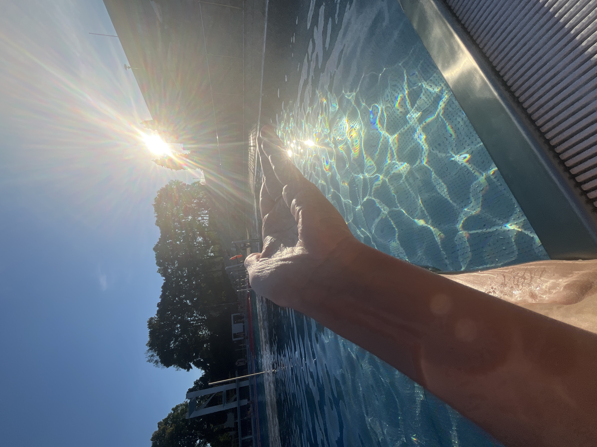 In an outdoor swimming pool, a person's outstretched hand is shown from a first-person perspective. The hand is cupped, with sunlight shining through it, casting rainbow-like refractions on the water's surface. The pool water is a clear blue, and the bottom is visible. Sunlight beams dramatically from the upper right corner of the image, indicating it is either sunrise or sunset. Trees and a building with a diving board are visible in the background, suggesting the location is a public swimming facility. The deck of the pool is visible in the foreground, with a grey, ribbed texture.