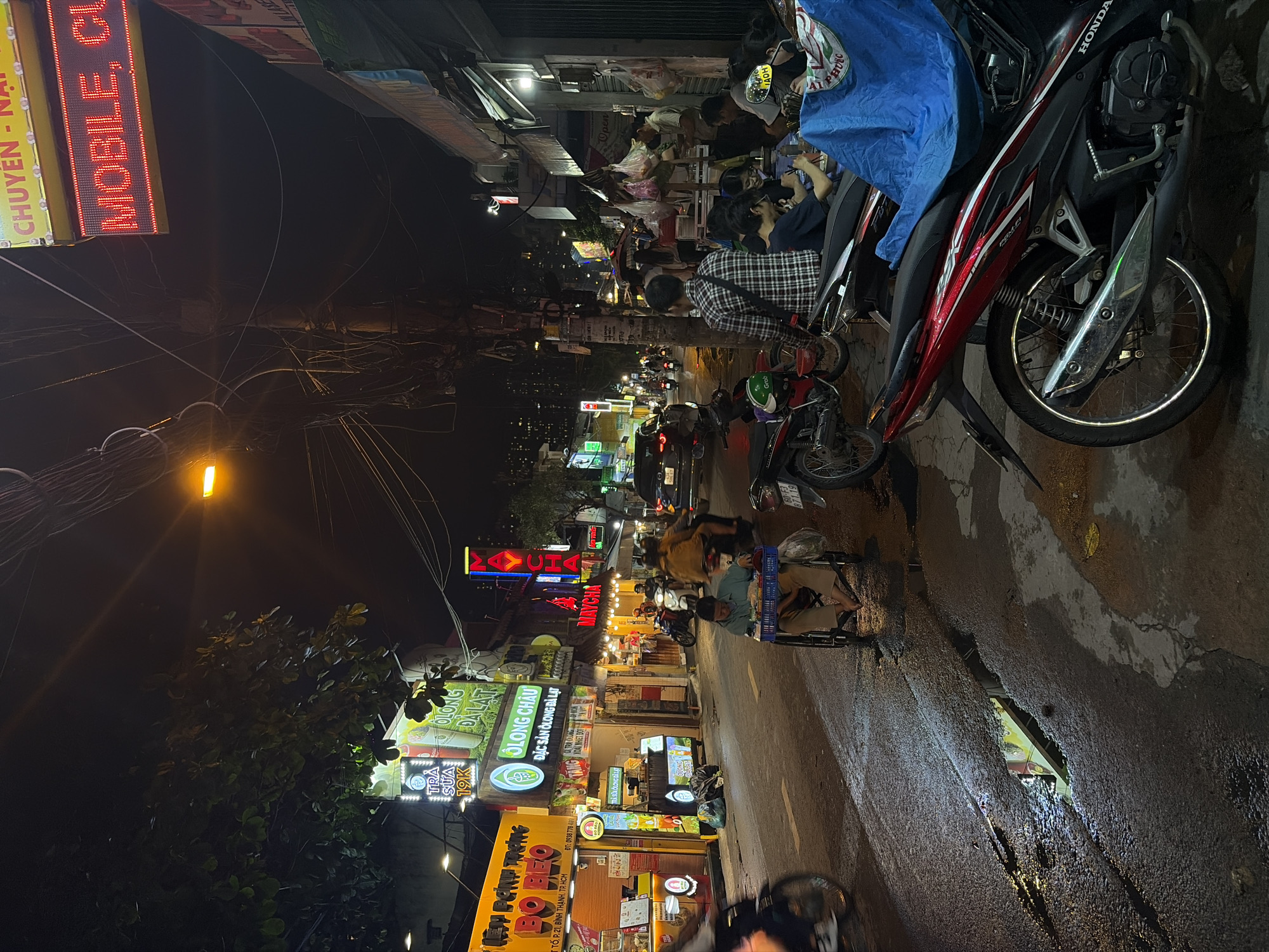 This detailed image captures a nighttime street scene in Thủ Đức, Ho Chi Minh City, Vietnam, characterized by vibrant commercial activity and urban infrastructure. The asphalt road, appearing wet in sections, reflects the numerous neon and LED signs illuminating the scene.
On the left side of the street, multiple storefronts display brightly lit signs, including "BÙ BÉO," "OLONG ĐÀ LẠT," and "TRÀ SỮA 19K," indicating food and beverage establishments. One sign explicitly displays "THỦ ĐỨC, HỒ CHÍ MINH," confirming the specific location. A barefoot individual propels themselves forward in a wheeled seat, carrying a blue plastic crate on their lap, positioned near the center of the wet road.
Traffic consists primarily of motorbikes, with several parked along the right sidewalk and others in motion, including a person riding a motorbike with a passenger further down the street. A black sedan is visible driving away in the middle distance. Overhead, a dense network of utility wires spans across the street.
On the right side, a street food area is active, with several individuals seated on low stools, appearing to be dining or socializing. A prominent red Honda motorbike is parked in the foreground, partially obscured by a blue tarp draped over its rear section. Above, a red LED sign displays "MOBILE, CU" and "CHUYÊN - NẠP," suggesting a mobile phone or charging service. Puddles on the pavement reflect the overhead streetlights and shopfront illumination. Dark foliage from trees is visible against the night sky on the left.