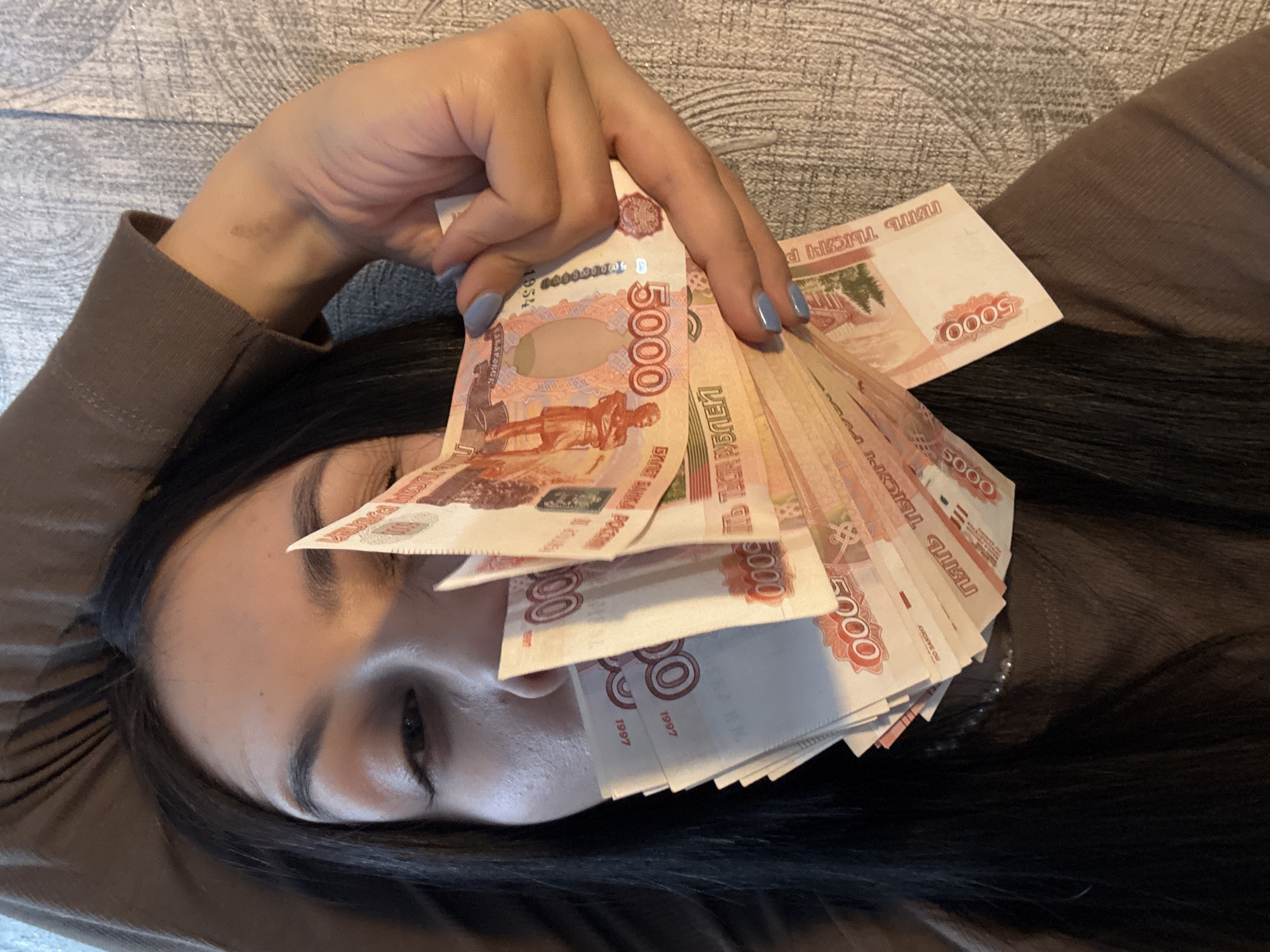 A woman lies on her back, facing upward, with a fan of Russian 5000 ruble banknotes obscuring part of her face. Her dark hair frames her face and shoulders, and she wears a brown long-sleeved shirt. Her nails are painted a light blue. The background appears to be a textured gray surface, possibly a couch or bed. The banknotes are prominently displayed, with "5000" visible on each. The city of Votkinsk, Russia is not explicitly visible or indicated in the image.