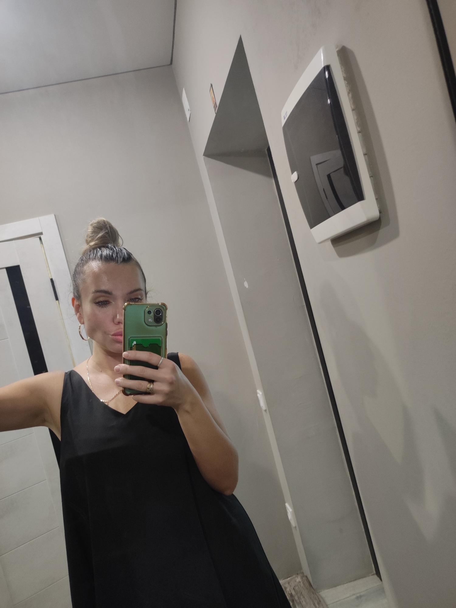A woman is taking a selfie in front of a mirror. She has her hair in a bun, and is wearing a black top. She is holding a green phone in her hand. The background includes a door, a wall, and a dispenser. The location is Votkinsk, Russia.