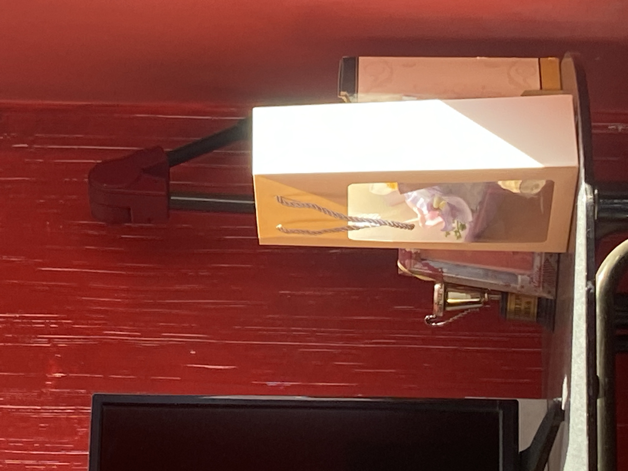 The image displays an arrangement of objects against a striking red wall, possibly located in Troyan, Bulgaria. The wall features a painted or textured surface with numerous horizontal light streaks, creating a distressed or wood-grain effect.
Centrally placed is a light brown gift box with a white top section and a clear display window. Dark, braided handles are visible on the box, which contains soft, pastel-colored items, likely artificial flowers or decorative elements in shades of light purple and white. A strong, diagonal shadow from an overhead light source, possibly sunlight, falls across the white top of the box.
To the left, a red clamp, resembling a spring clamp, is attached to the red wall, securing a dark bar or rod. Directly below the gift box, a small, polished brass-colored bell with a chain hangs from an obscured structure. Part of a metallic, vertical frame or support structure is visible along the right edge of the image. At the bottom of the frame, the top portion of a black electronic screen, such as a computer monitor or television, is present.
No people or direct activity are depicted; the scene is a static composition of these items. The bright, directional lighting indicates it is daytime. While some faint patterns are visible on packaging above the gift box and on the object from which the bell hangs, no discernible text is readable.