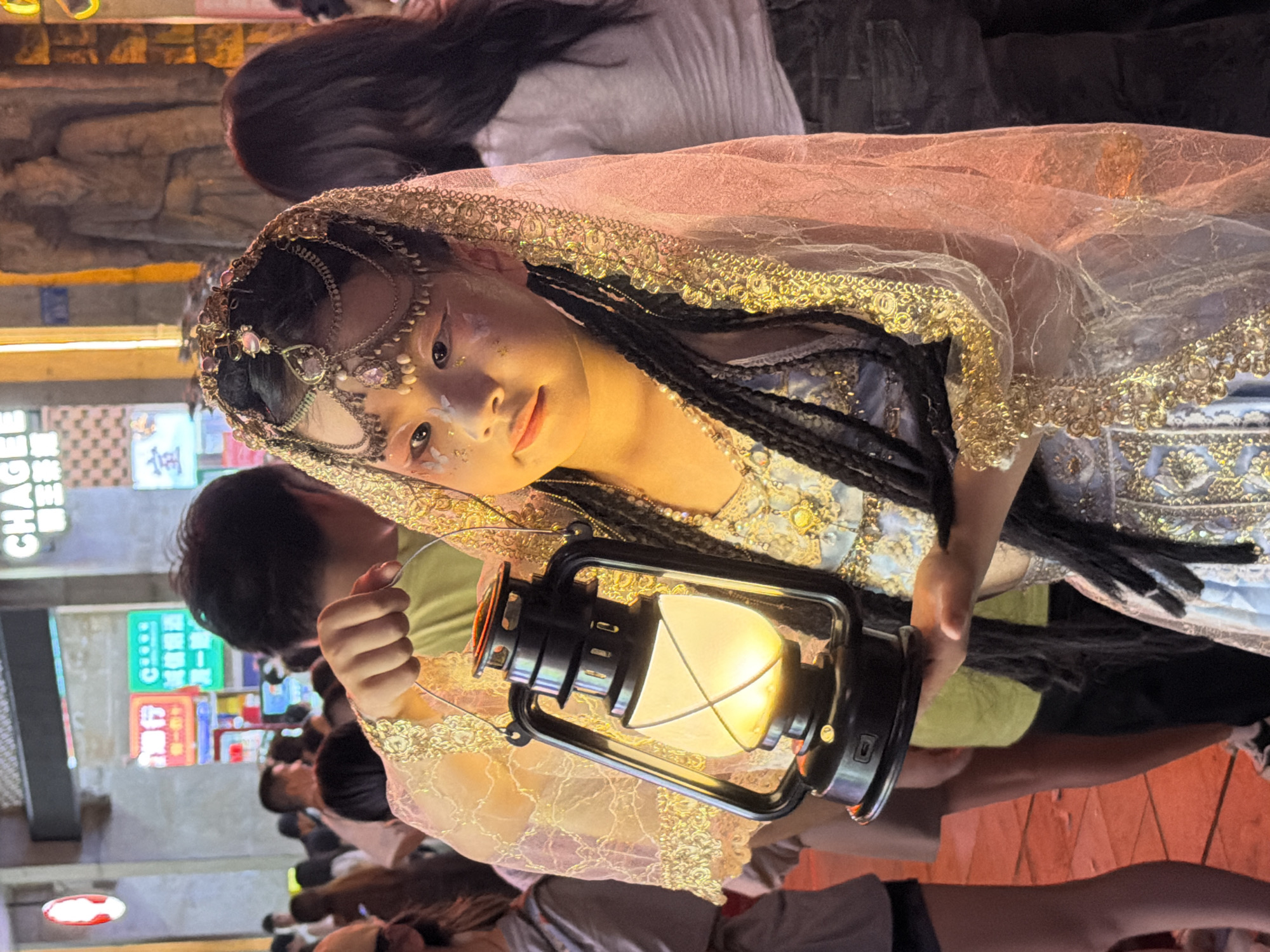 A person, likely a young woman, is depicted in the foreground, holding a black lantern that emits a warm yellow glow. She is adorned in an elaborate costume, featuring a sheer veil with gold trim and intricate beadwork on her head. Her makeup is striking, with white accents and glitter around her eyes. Her dark hair is styled in braids. The subject is looking towards the camera with a gentle expression.
The scene appears to be outdoors, possibly at night, given the artificial light of the lantern and the illuminated signs in the background. The background reveals a bustling street scene with various people and colorful signs, suggesting a public gathering or event. The signs in the background feature Chinese characters. One sign clearly reads "CHAGEE" in English, with Chinese characters above it. Another sign has green text on a white background. The overall atmosphere is festive and vibrant, typical of a night market or cultural festival in Yuzhong District, China. The lighting suggests it is evening or night.