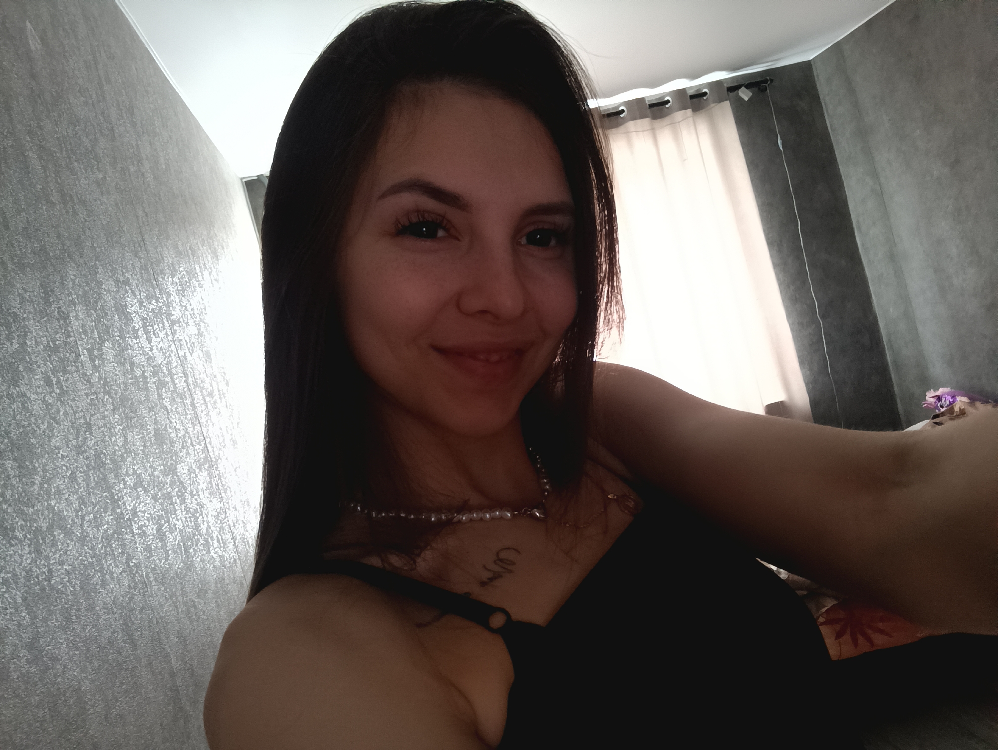 A woman with dark hair and a pearl necklace is facing the camera and smiling slightly. She is wearing a black top with thin straps. A small tattoo is visible on her chest. The background includes a gray wall on the left and a window with white curtains on the right. The location is an interior space within a city of Unknown, Unknown.
