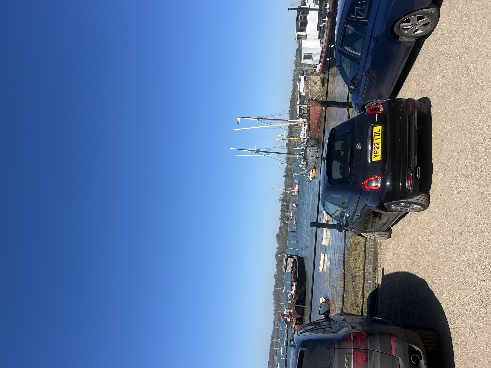 The scene shows a waterfront area in Babergh, United Kingdom, on a clear day with a cloudless blue sky. Three cars are parked along a waterfront street. A black Fiat 500 with license plate YP22 VDL is parked between two larger, darker cars. The Fiat is positioned directly next to a low fence that runs along the water's edge. Beyond the fence, a body of water (likely the River Stour) is visible with several boats moored. Some have masts. The edge of a building is visible on the far side of the water.
