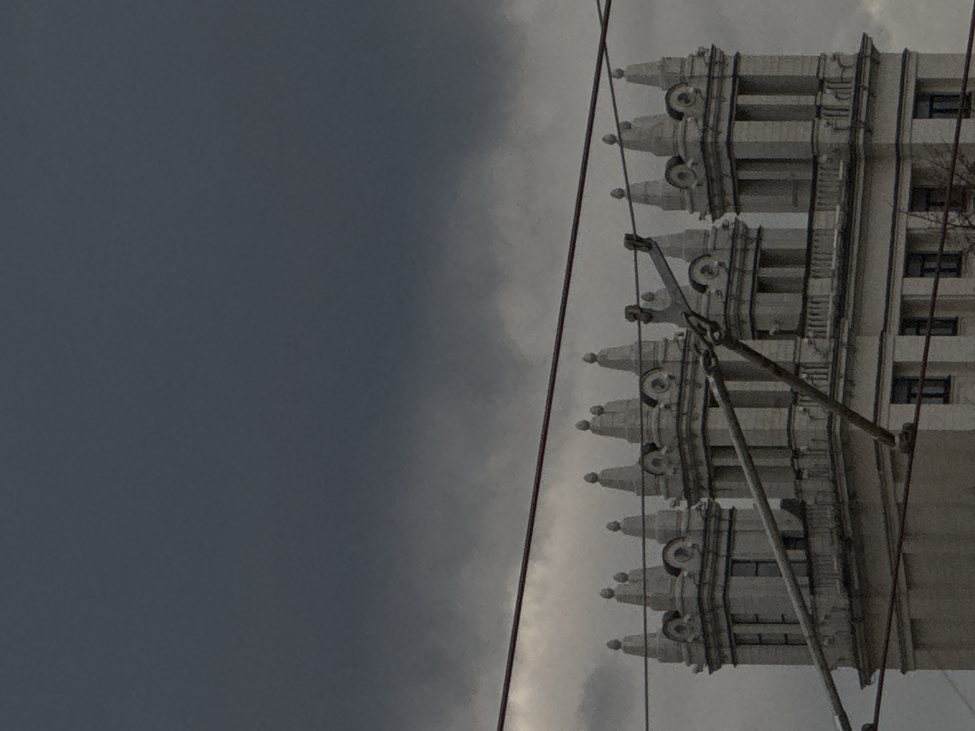 The image shows a tall, grand building under an overcast sky, with utility lines crossing the foreground. The building occupies the right half of the frame, characterized by its ornate, monumental architecture, likely in the Stalinist Empire style, which aligns with the provided context of Moscow, Russia.
The building features multiple levels of decorative elements along its upper facade, including repeated spires or finials and corbels. These architectural details create a sense of grandeur and historical significance. Numerous windows, appearing dark, are visible across the building's face. The structure is rendered in a light, possibly grey or off-white, material, contrasting subtly with the darker sky.
The left half of the image is dominated by a heavily clouded, grey sky, indicating an overcast day. No direct sunlight is visible, contributing to a muted and slightly somber atmosphere. Running diagonally across the upper-right portion of the image, and again in the lower-left, are dark utility lines, likely power or tram lines, with visible insulators and supports. These lines partially obscure the building.
There are no people or visible text in the image. The scene captures a static architectural view on a cloudy day in an urban setting.