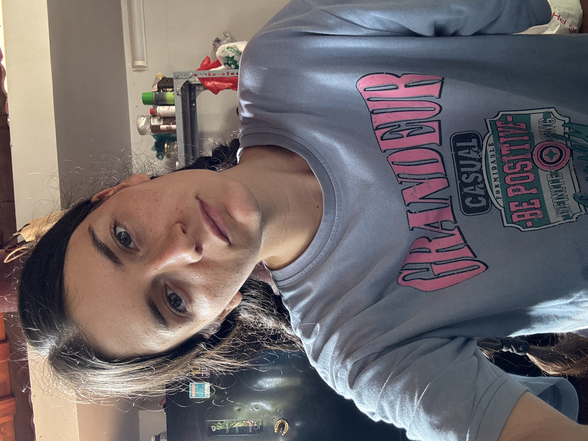 A woman with dark hair, styled in pigtails, faces the camera while wearing a light blue shirt with the words "GRANDEUR CASUAL" and "BE POSITIVE INFINITE" printed on it. The background shows a kitchen setting with shelves containing jars and containers. A refrigerator with magnets on the door is also visible. The location of the image in Mũi Né, Vietnam, cannot be confirmed from this image.