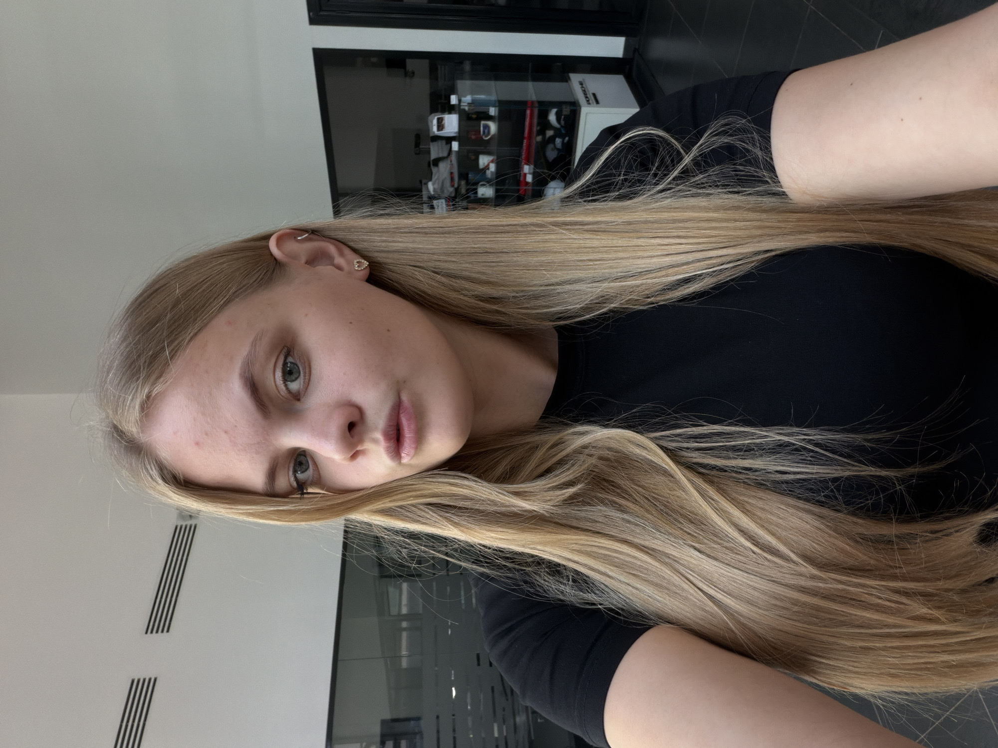 A woman with long blonde hair and fair skin is pictured indoors. She is wearing a black t-shirt and earrings, with the left ear showing a small hoop earring and a small stud. Her gaze is slightly upward. Behind her is a white wall with black linear decorations and what appears to be a glass wall with a storage unit filled with various objects visible through it. The location in the city of Izhevsk, Russia, is undetermined from this image.