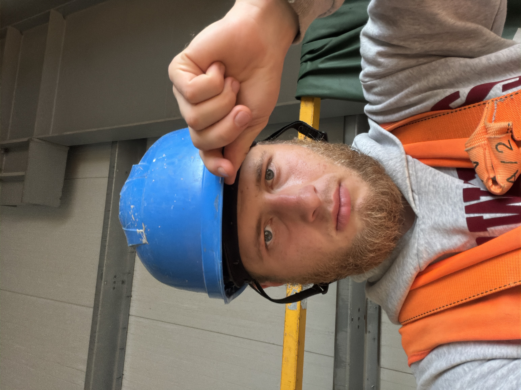 The image shows a close-up, upside-down portrait of a young man wearing a blue hard hat and an orange safety harness over a grey hoodie. He has light skin, light eyes, and a reddish-brown beard. His right hand is held up, with his fist near the top of the frame, touching the brim of his hard hat. He is looking directly at the camera with a neutral expression.
The background suggests an industrial or construction setting. There are metal beams and grey paneling visible, possibly part of a building under construction or a warehouse. A yellow support beam is also visible behind the man's head. The lighting appears to be artificial, suggesting it could be indoors or on a cloudy day.
The orange safety harness has a label with text that is partially visible. It appears to read "1/2" or "12".
The context provided indicates the location is Stróża, Poland, which further supports the interpretation of an industrial or construction environment. No specific time of day or weather conditions are discernible from the image itself. The overall mood is utilitarian, focusing on the worker and his safety gear.