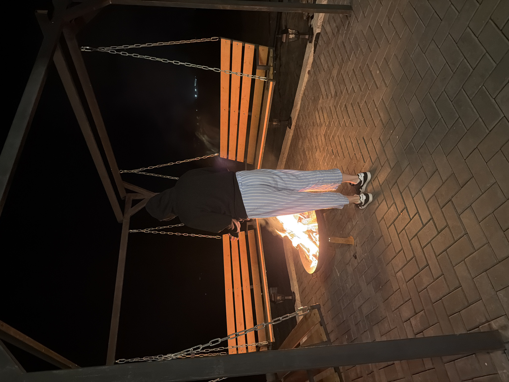 A hooded person wearing striped pants and sneakers stands in front of a fire pit situated near a wooden swing. The surrounding ground is paved with brick. The scene appears to be at night. The location is likely Kamennoye, Russia.