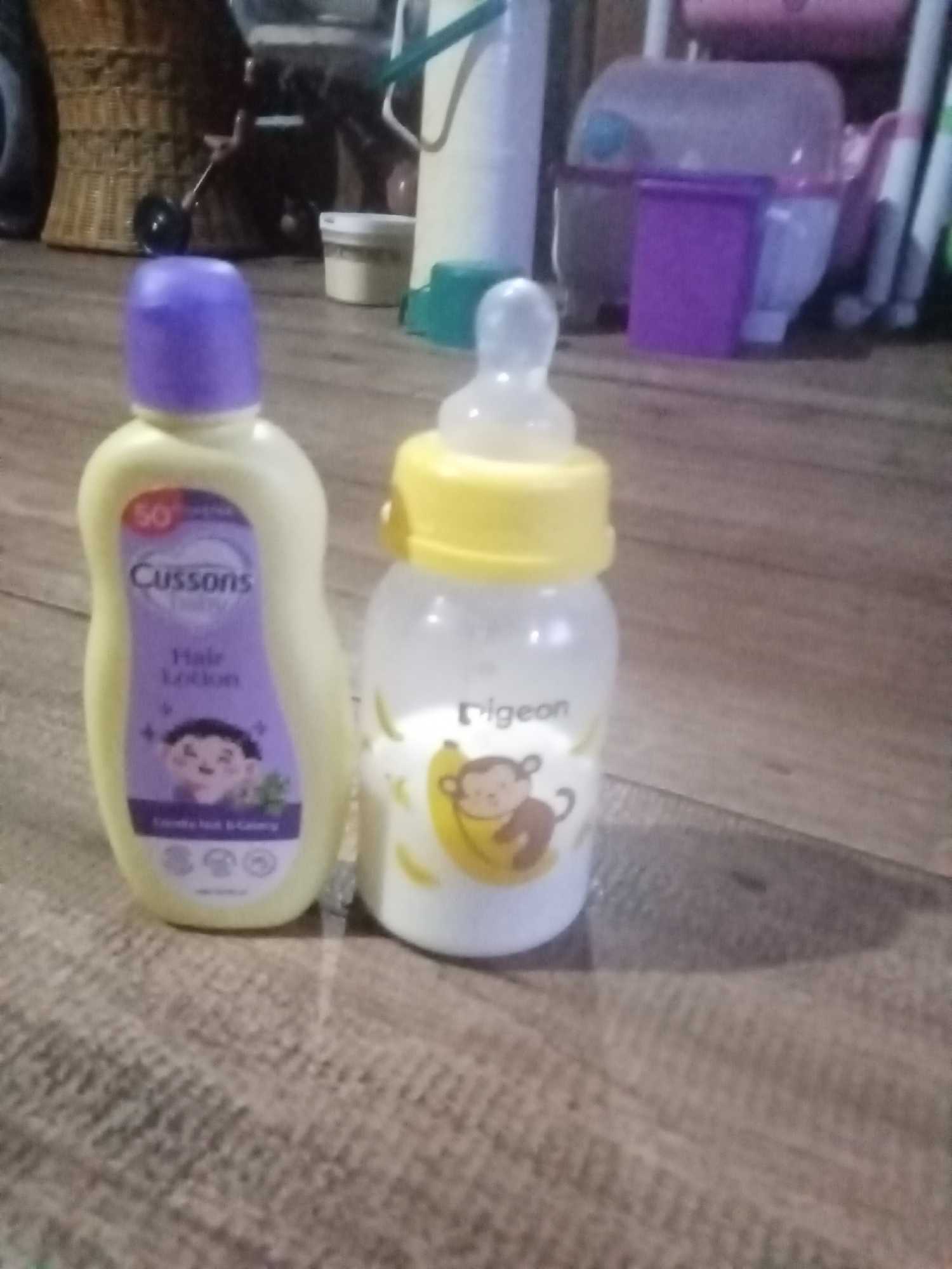 In the city of Setia Jaya, Indonesia, a close-up, slightly blurred image captures two baby care items on a wooden surface. On the left is a light yellow bottle with a purple cap, identified as Cussons Baby Hair Lotion. The bottle has a purple label featuring a smiling baby illustration and text. To its right is a clear baby bottle with a yellow nipple and cap, containing a white liquid. The bottle has a label with a cartoon monkey holding a banana and the brand name "pigeon." The background is out of focus, showing various household items and textures, including what appears to be wicker furniture and plastic containers.