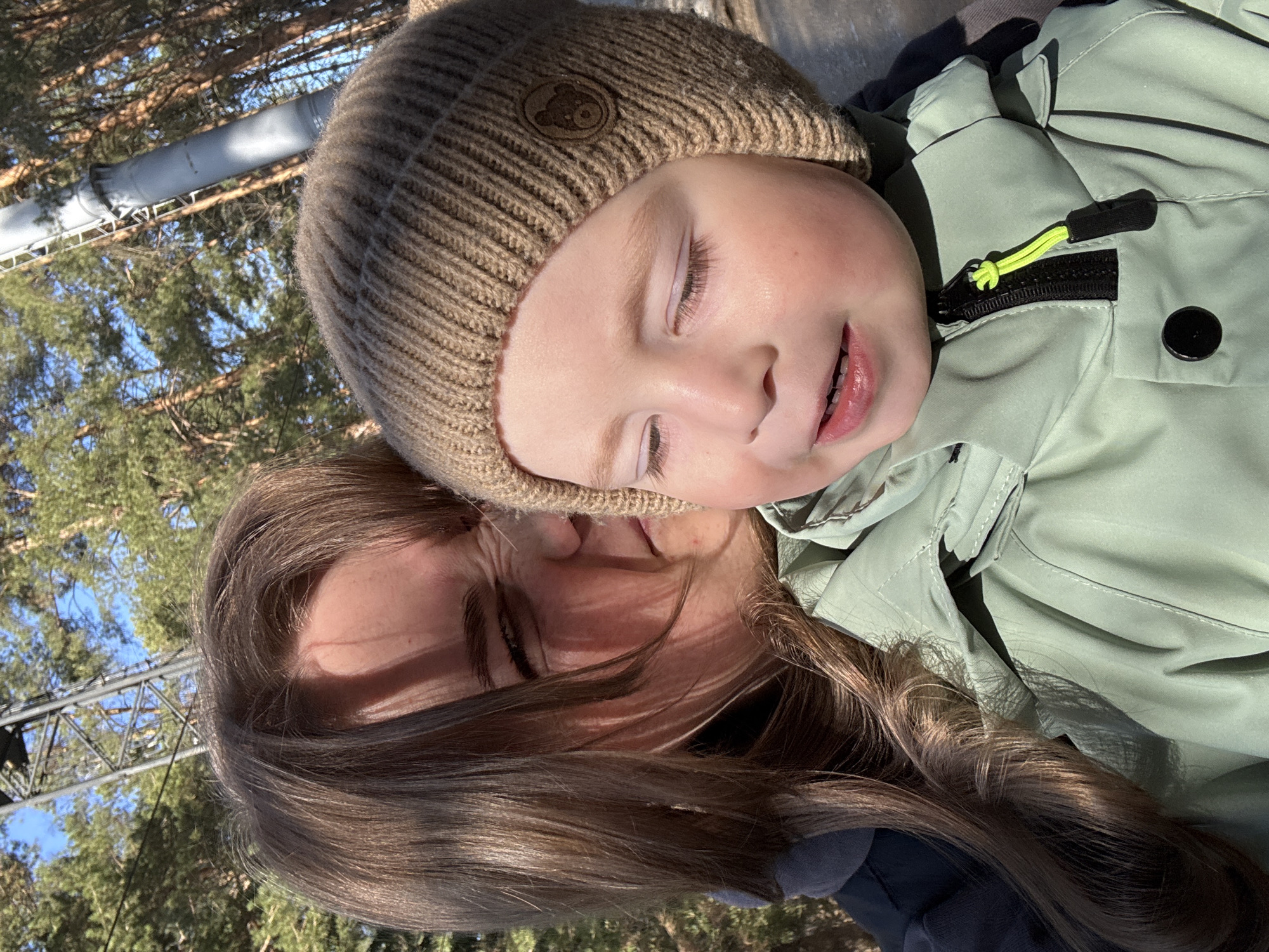 A woman with long brown hair holds a young child in her arms. The child wears a brown knit hat with a bear emblem and a light green jacket with a bright yellow zipper pull. Both have their eyes closed and are facing towards the viewer. The background shows trees and a metal structure suggestive of a ski lift. The location is likely in the city of Chaikovsky, Russia, a region known for outdoor recreational activities like skiing.