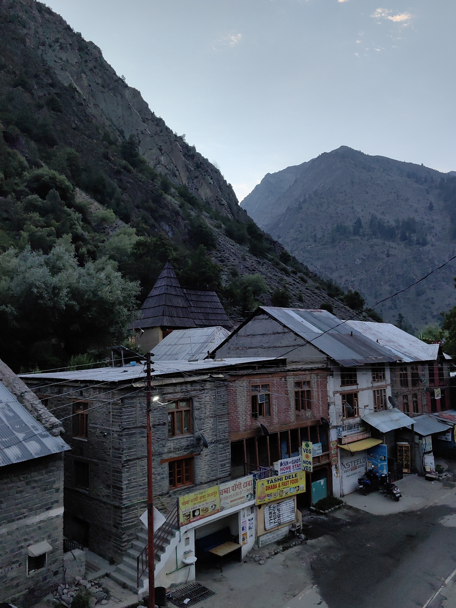 The image depicts a mountainous landscape with a village nestled at its base. In the foreground, there are several multi-story buildings constructed with stone and brick. Some of these buildings appear to be shops or guesthouses, indicated by signs with text in what appears to be Hindi. One sign reads "TASHI DELE DHABA & FAST FOOD." The roofs are a mixture of corrugated metal and what looks like slate tiles. A paved road runs in front of these buildings, with a few motorcycles parked on the side. A red utility pole with streetlights stands prominently in front of one of the stone buildings. The background features a steep, rocky mountain covered with sparse vegetation and trees, under a pale sky. The overall impression is of a remote, possibly underdeveloped settlement in a rugged, natural environment. The city of Udaipur, India is mentioned as the location, but the scene itself does not contain any specific landmarks that would immediately identify it as Udaipur. The image appears to be taken from a slightly elevated vantage point, possibly another building or a higher ground.