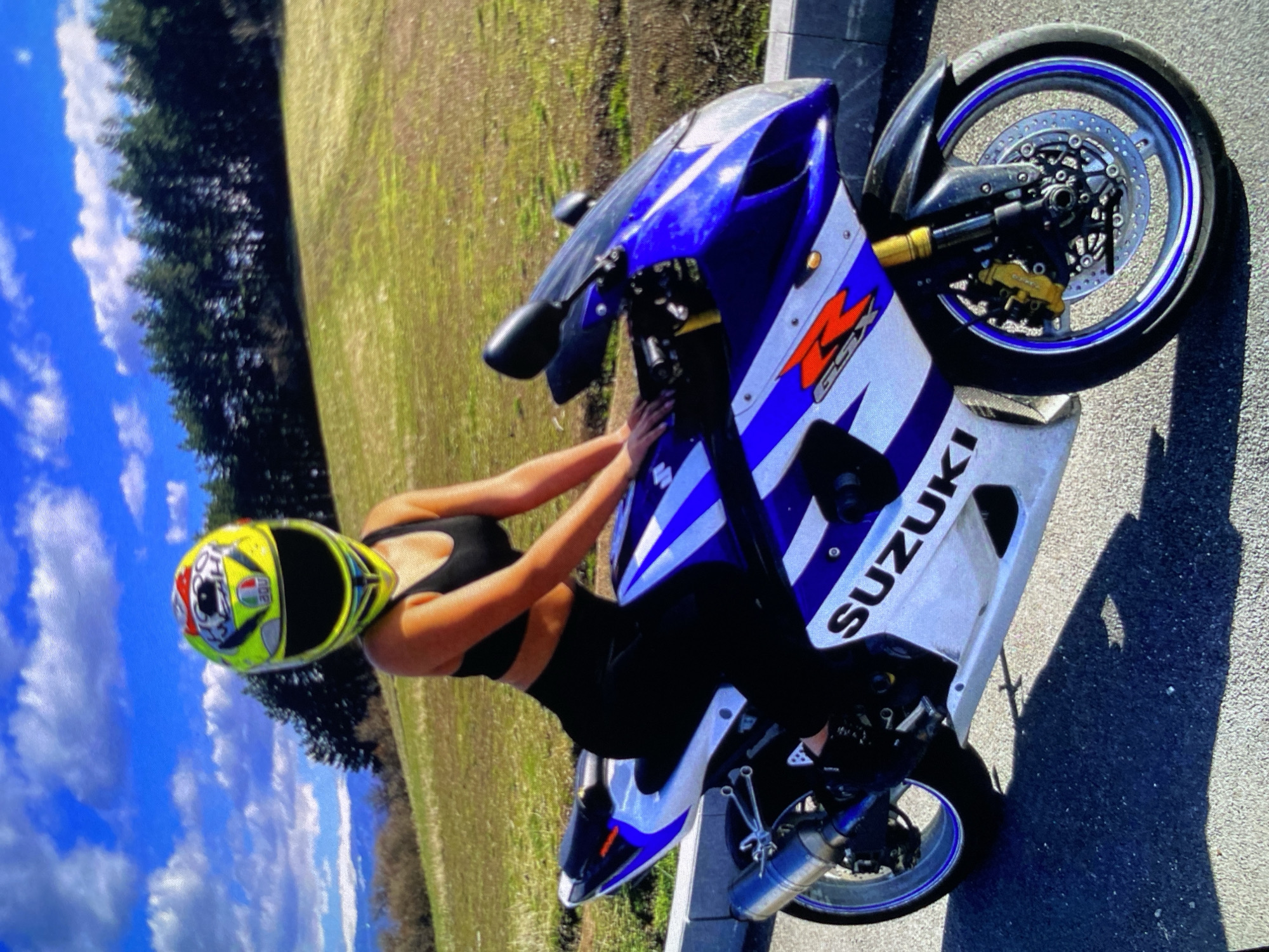 An outdoor, eye-level shot captures a person posing with a blue and white Suzuki sport motorcycle on a sunny day in Bankya, Bulgaria. The image is oriented sideways, with the ground appearing on the right and the sky on the left.
The main subject is a person, likely female, wearing a black crop top and black pants. They are also wearing a brightly colored yellow, red, and white full-face helmet with "AGV" text and intricate graphics visible. The person is positioned leaning over the tank of the blue and white Suzuki motorcycle, with their hands resting on the handlebars.
The motorcycle is a Suzuki GSX-R, indicated by the "SUZUKI" text prominently displayed in white letters on the blue and white fairing, and the red "GSX-R" logo near the front. It features a blue and white color scheme with white stripes on a blue background. The motorcycle has blue accents on its wheels and a silver-colored exhaust pipe. The front wheel shows detailed disc brakes.
The setting is outdoors under a clear blue sky with scattered white clouds. In the background, there is a lush green grassy field and a line of dark green trees. The motorcycle is parked on an asphalt surface next to a light-colored curb, suggesting a roadside location. The strong sunlight casts visible shadows on the asphalt. The time of day appears to be mid-day due to the bright illumination.