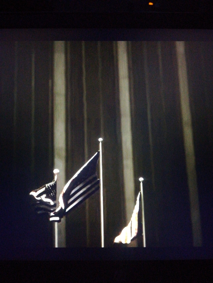 Three flagpoles display flags, two are American flags, and one is unidentifiable. The flags are illuminated from below, casting light onto the dark fabric and poles. Behind the flags, a pattern of dark and light vertical lines rises into the darkness. The image is dark and grainy.