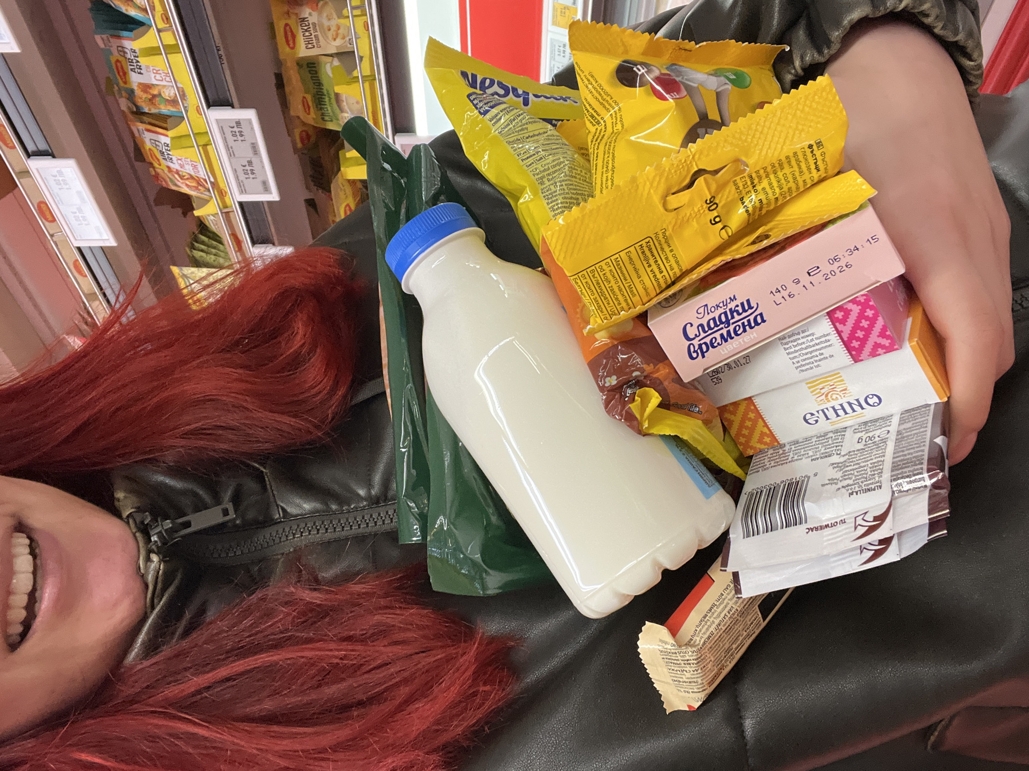 Here is a detailed description of the image:
**Scene:** The image appears to be taken inside a supermarket in Asenovgrad, Bulgaria. The person in the photo is holding a small assortment of groceries in their hands or a small shopping cart.
**People:** Only a portion of the person holding the items is visible. They have red, shoulder-length hair and are wearing a black leather jacket. Only the person's mouth and teeth are visible.
**Objects:** The items being held include:
* A plastic bottle of white liquid, possibly milk or yogurt, with a blue cap.
* Multiple yellow snack packages, appearing to be some type of candies or chocolates.
* A pink box of "Локум Сладкӣ времена Цветен" (Lokum Sladki Vremena Cveten), a type of Turkish delight. The packaging indicates an expiration date of L16.11.2026.
* A package of ETHNO chocolate.
* Other assorted small snack packages.
In the background, supermarket shelves are visible, stocked with various food products. Price tags are hanging on the shelves, indicating the price of some of the items.
**Actions:** The person is holding the items, presumably after selecting them from the shelves.
**Environment:** The setting is a typical supermarket aisle, evidenced by the shelving and product displays.